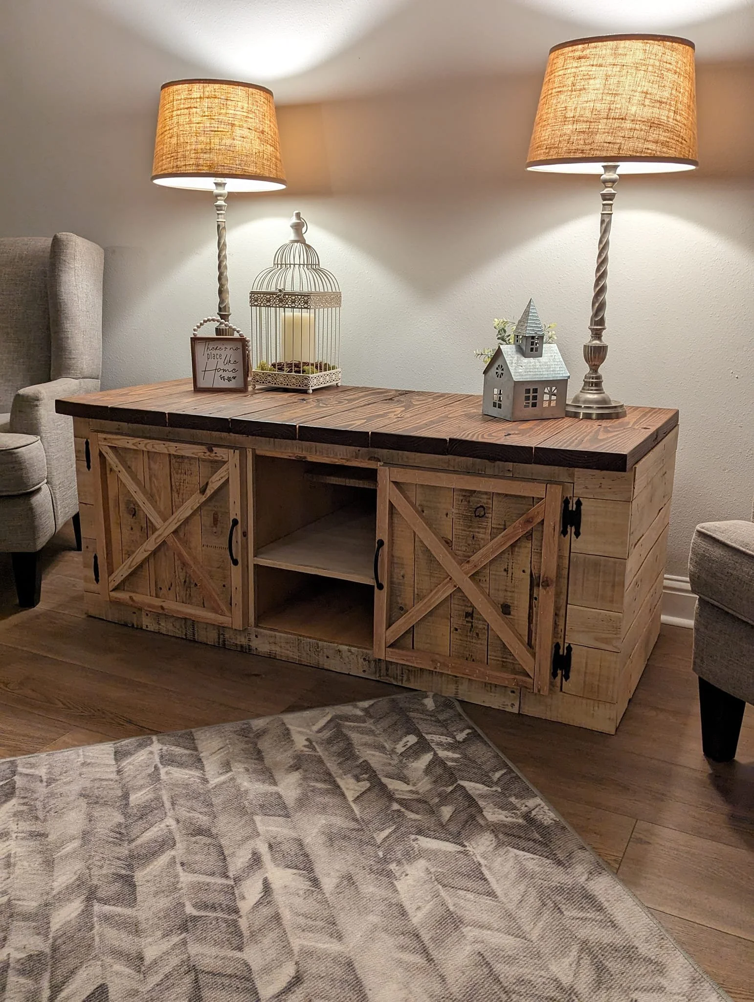 A wooden console table with decorative items on top, illuminated by two table lamps with beige shades, against a plain wall in a cozy living space.
