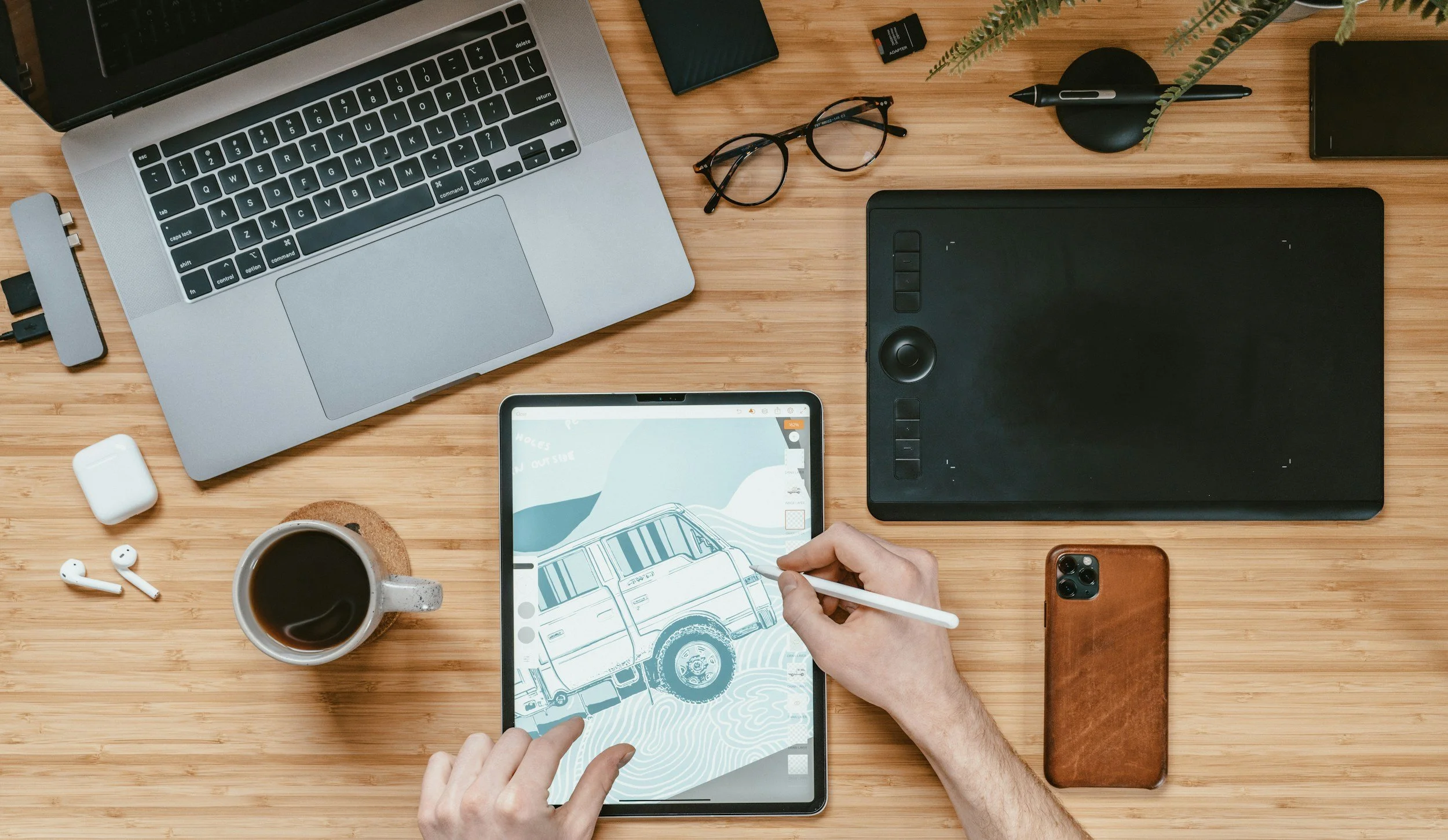 A wooden desk with a MacBook, glasses, a drawing tablet, a smartphone, earbuds, a coffee mug, a pen holder with a pen, an AirPods case, a SD card, an external hard drive, and a person drawing a car on a tablet with a stylus.