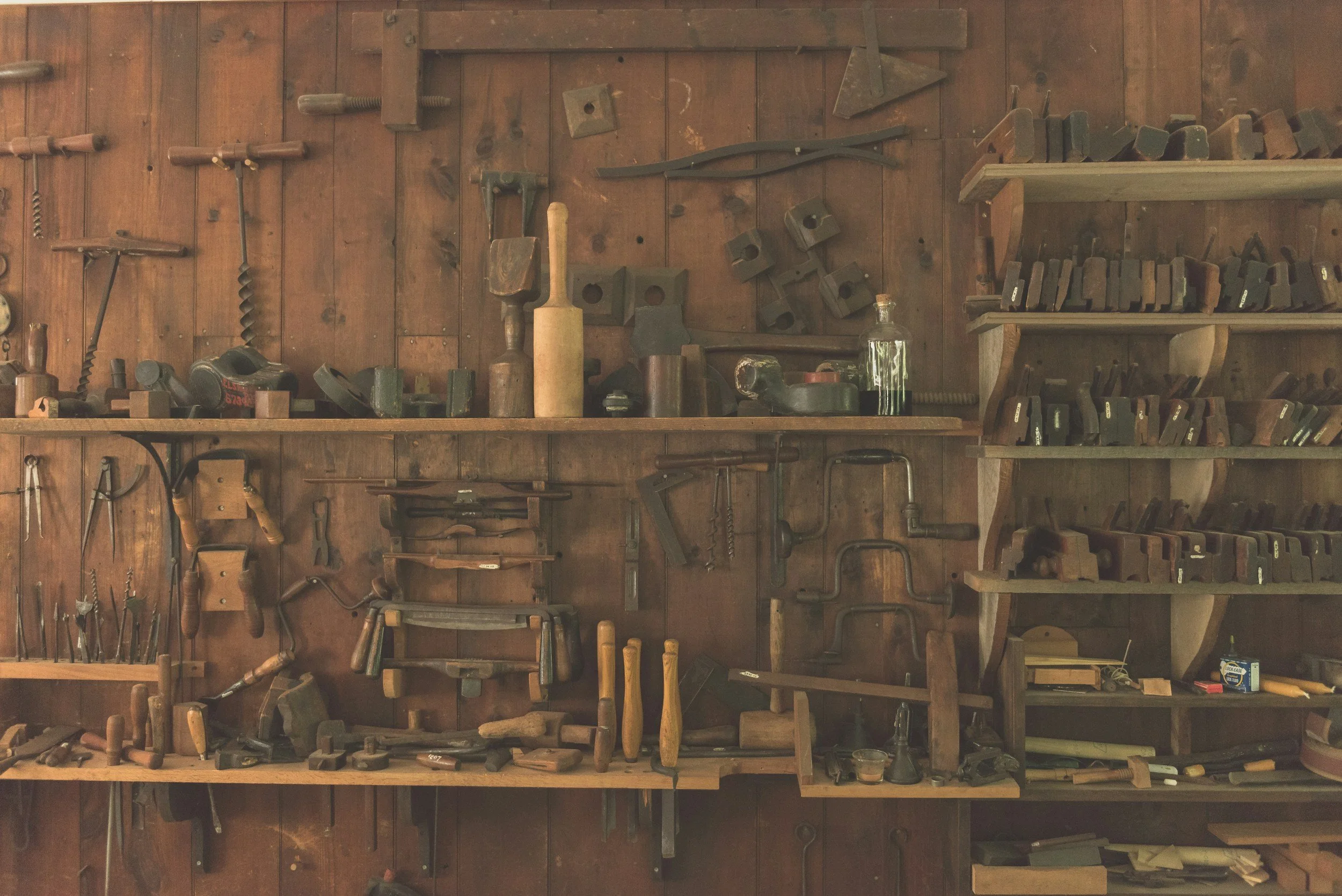 A wall with various old woodworking tools and equipment mounted on and organized on shelves, including hand planes, saws, clamps, and chisels.