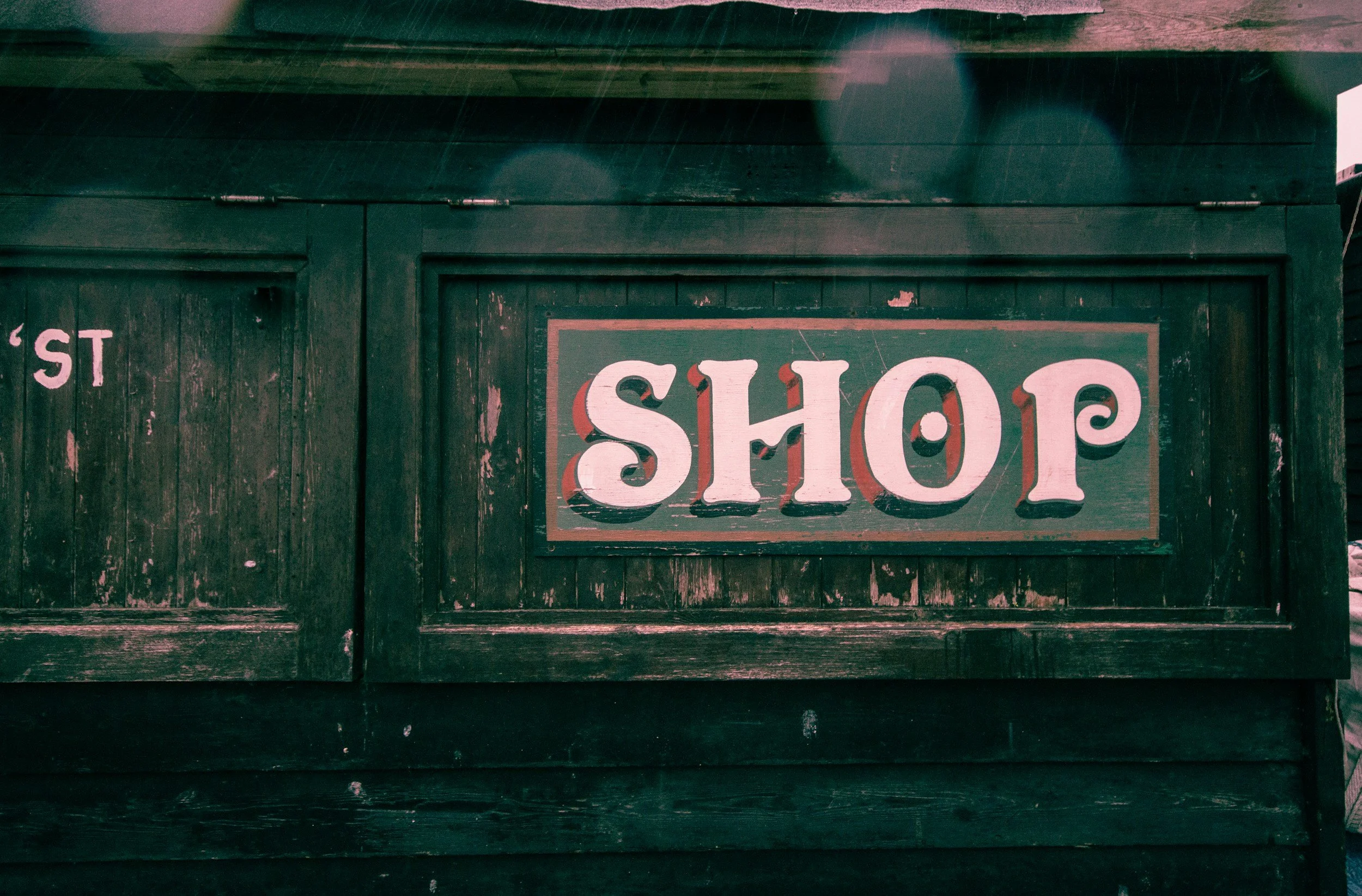 Close-up of a weathered green wooden shop sign with white and red lettering, partially visible, mounted on a dark painted wooden wall.