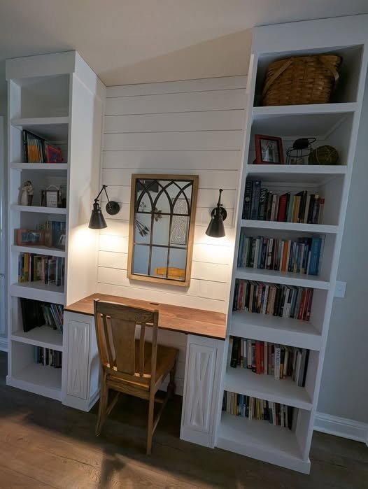 Home office with built-in white bookshelves, a wooden desk, a chair, a mirror, and wall-mounted lamps.