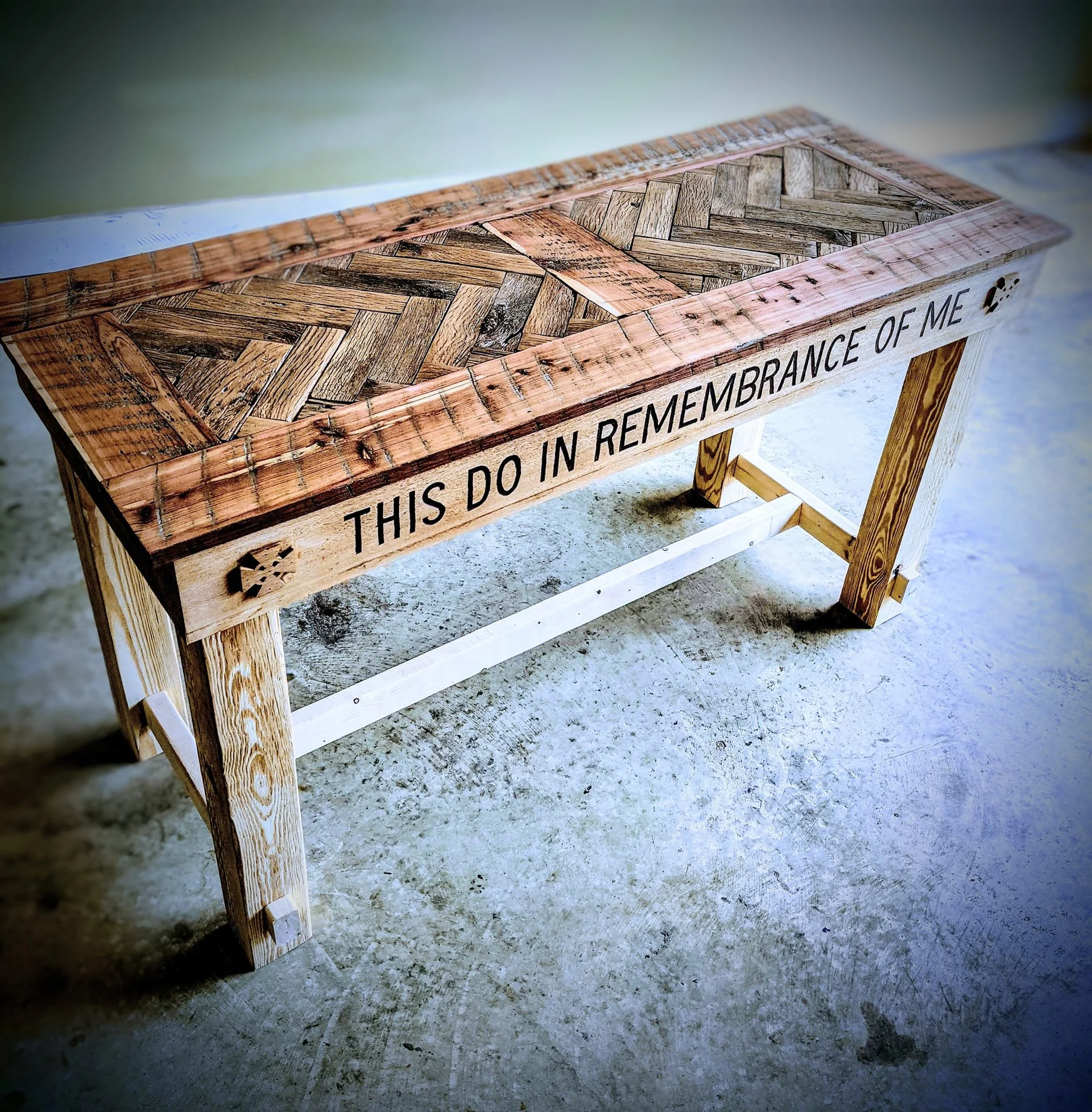 A wooden table with a herringbone pattern on its surface, placed on a concrete ground. The side of the table has a phrase written on it, partially visible, that reads 'This do in remembrance of me'.
