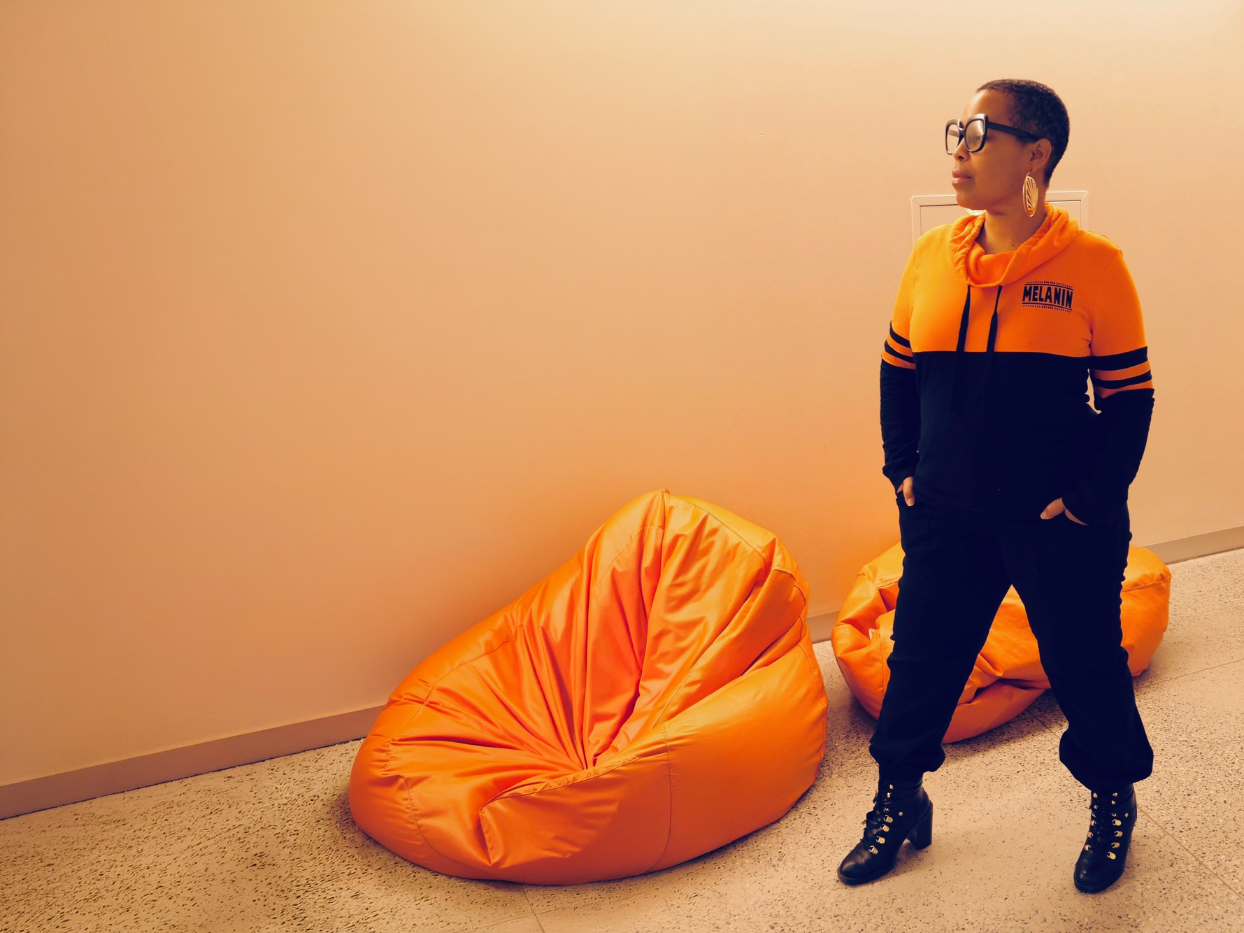 Jaz, with short hair, glasses, and large earrings stands in a room in an orange jacket. She is wearing a black and orange hoodie with the word 'MELANIN' on it, black pants, and black boots, looking to her left with her hands in her pockets.