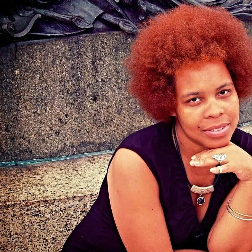 Jaz with short, curly reddish hair sitting outdoors near a textured stone wall, smiling slightly, wearing a dark sleeveless top and jewelry including rings, bracelets, and a necklace.