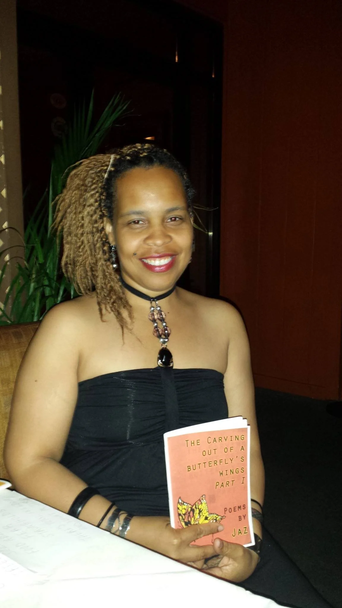 Jaz with dark and blonde dreadlocked hair, wearing a black strapless dress and jewelry, holding a book titled 'The Carving Out of a Butterfly's Wings Part I' by Jaz, sitting indoors.