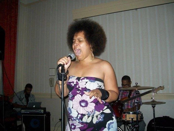 Jaz singing passionately into a microphone while performing with a band, including a guitarist and drummer, in an indoor setting.