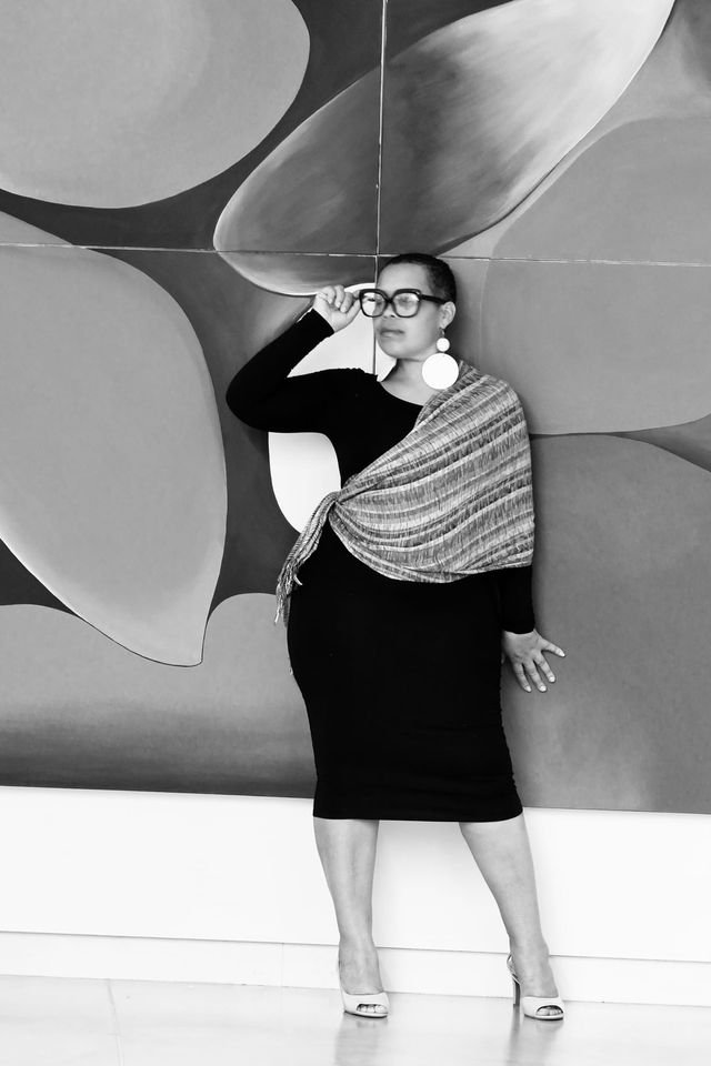 Jaz in a black dress and high heels, wearing large earrings and glasses, stands in front of an abstract mural. She holds one hand near her glasses and the other resting on the wall.