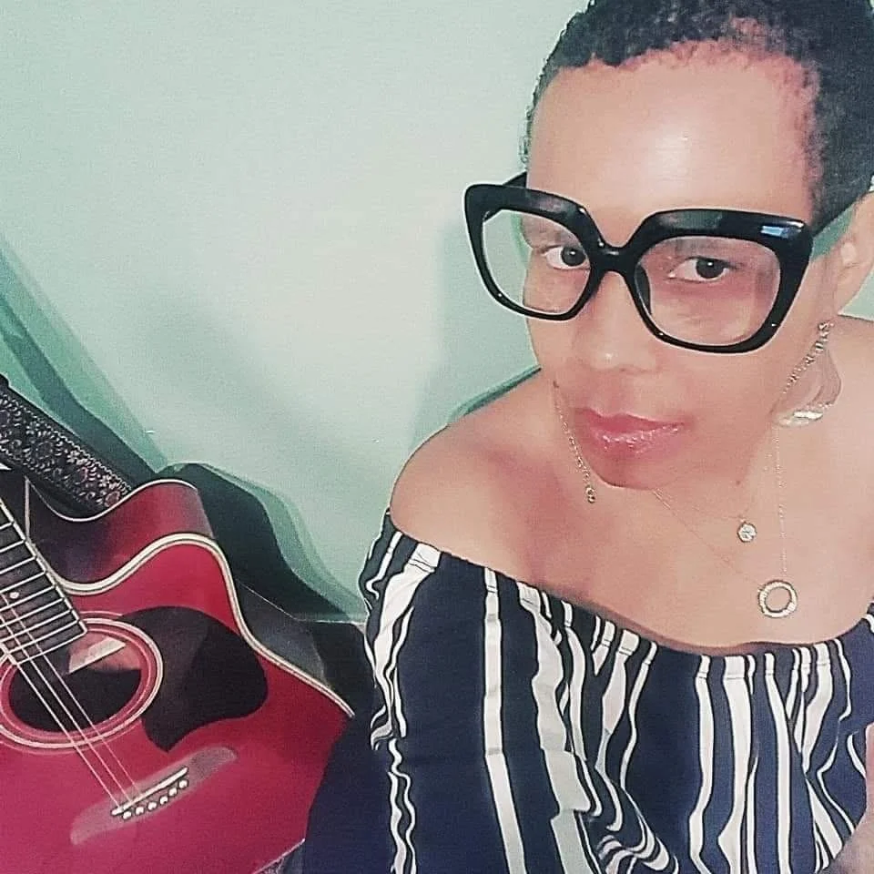 Jaz wearing large black glasses, off-the-shoulder striped top, and jewelry, sitting near a red acoustic guitar.