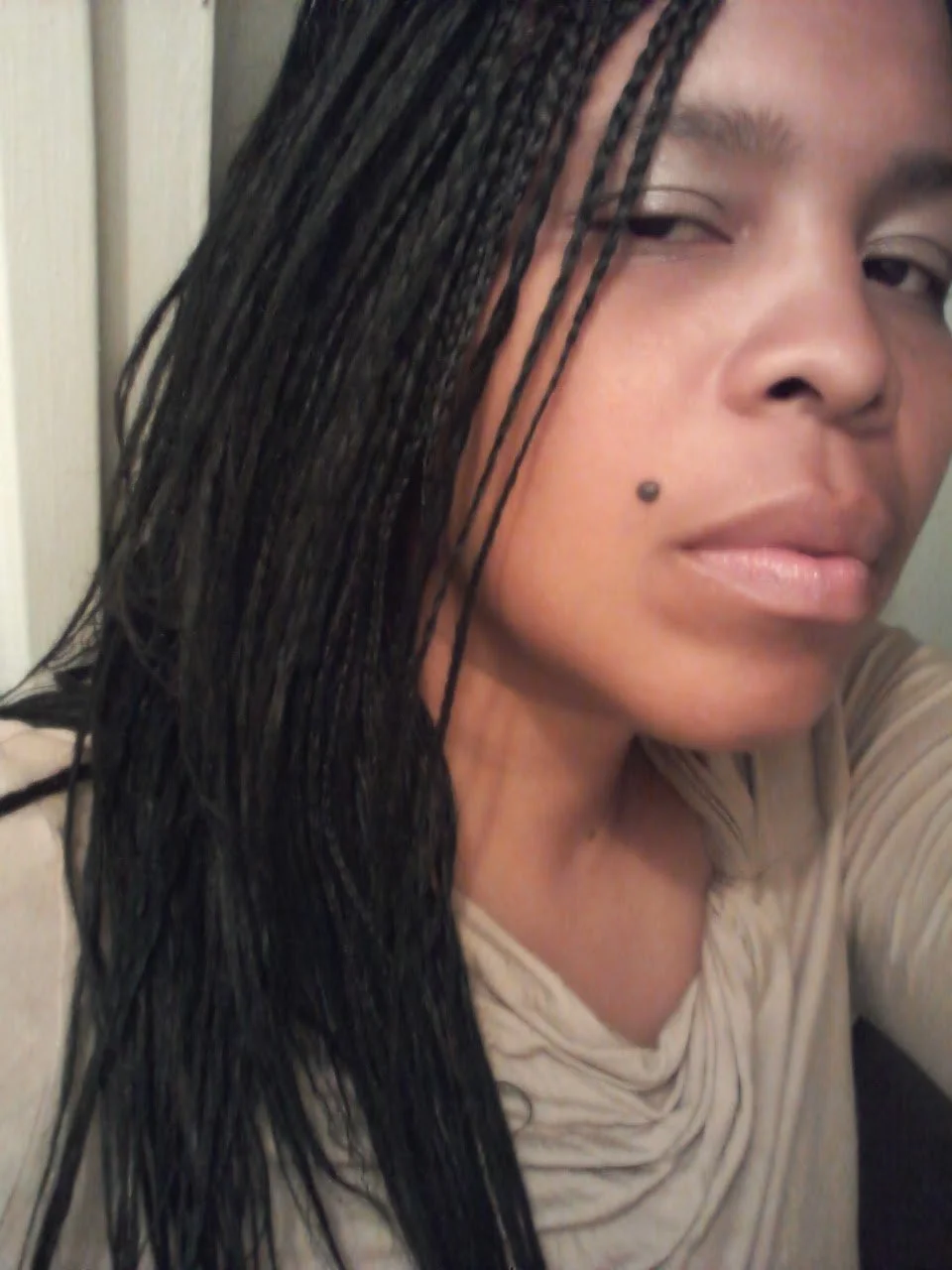 Close-up of Jaz with long black braided hair, light brown skin, a beauty mark on her face, and a beige shirt, partially visible.