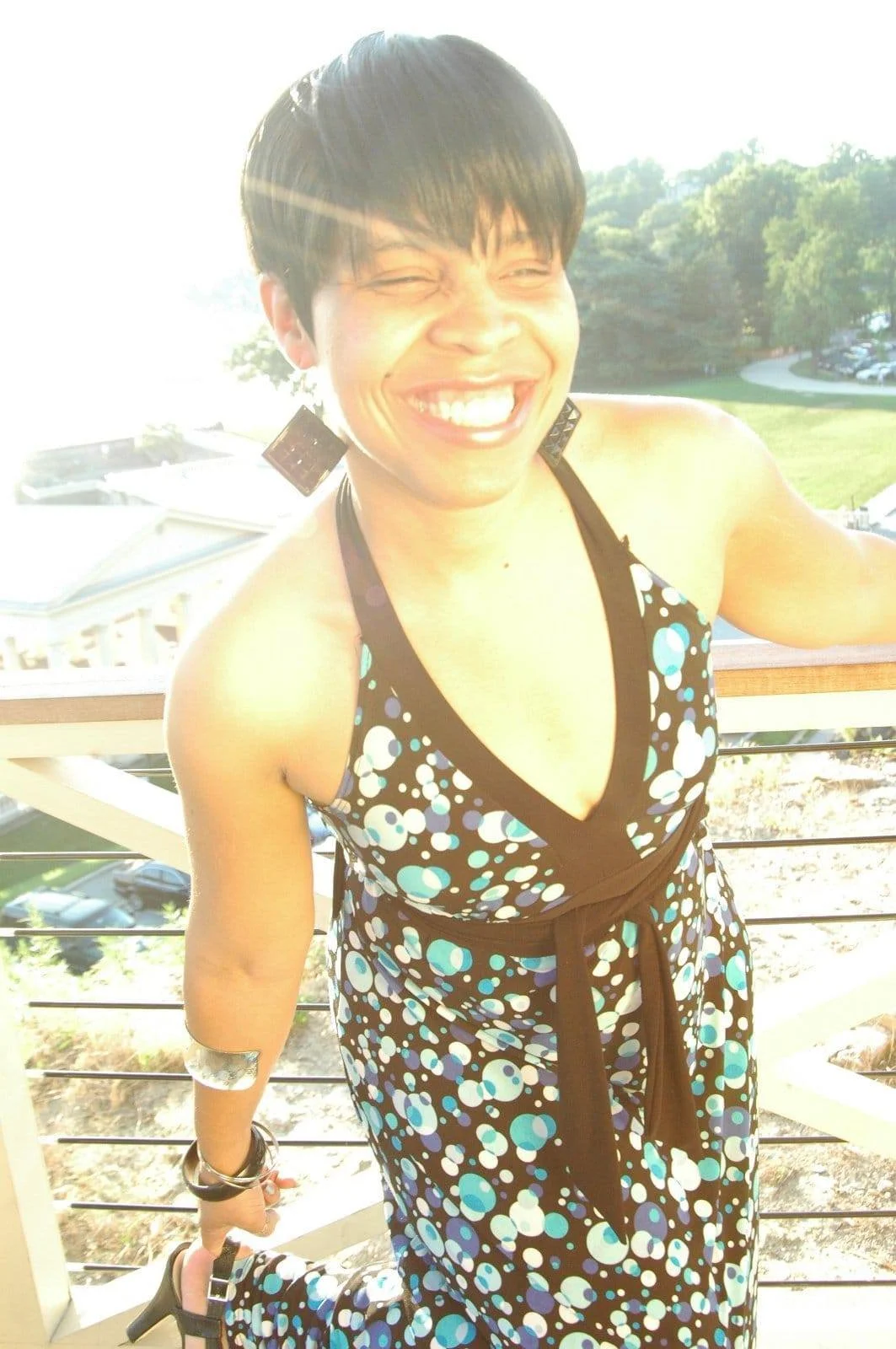 Jaz, with short black hair smiling broadly on a sunny outdoor balcony, wearing a black dress with blue and white polka dots, and large black earrings.