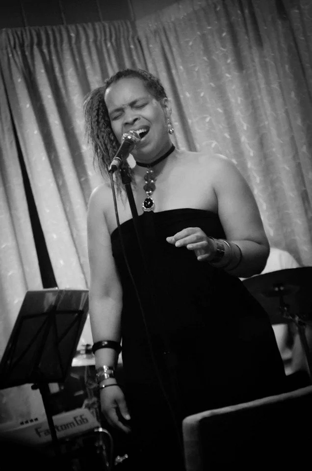 Jaz, singing passionately into a microphone on stage, wearing a strapless black dress and jewelry, with closed eyes and an expressive face.