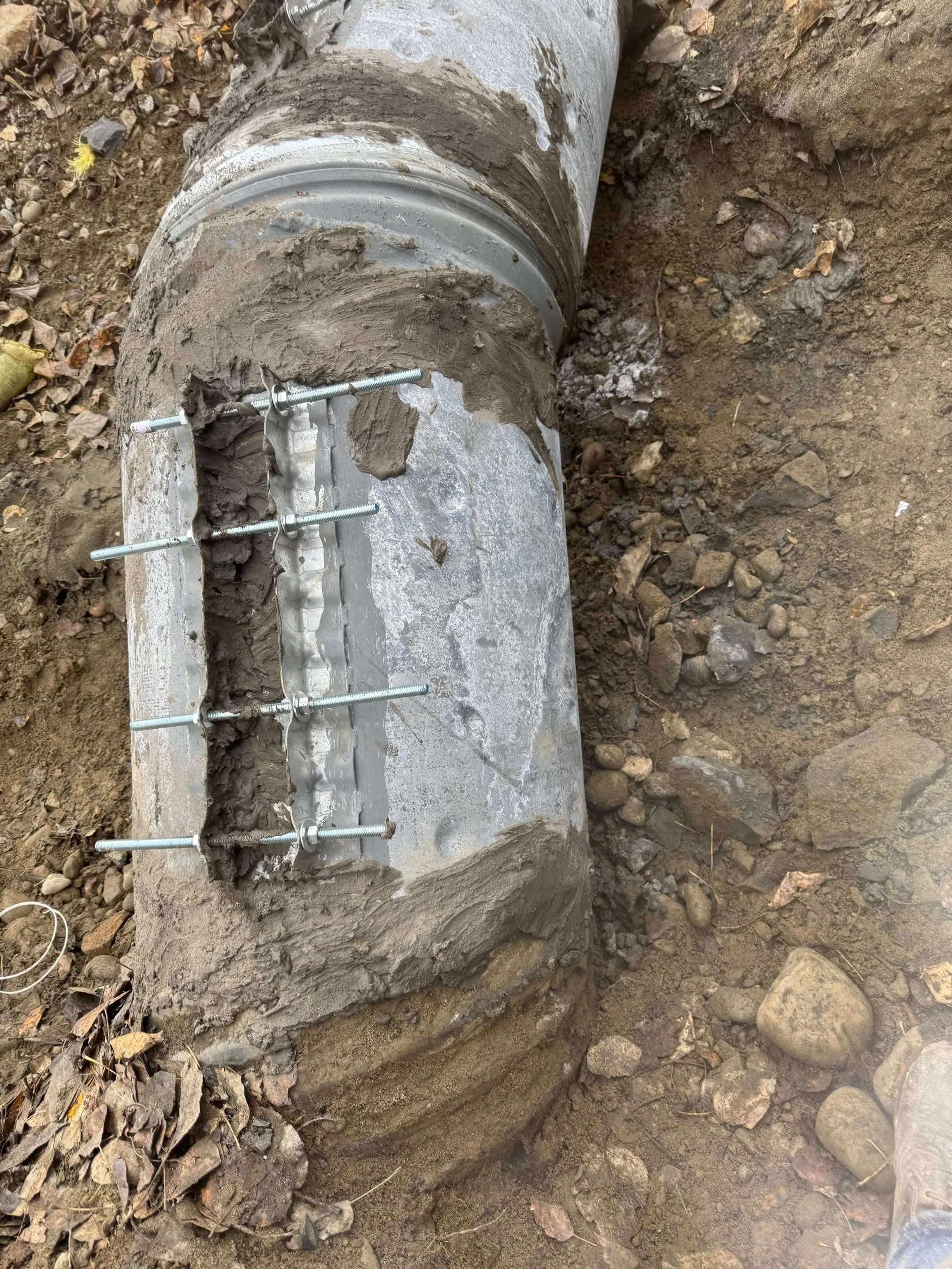 A large concrete pipe is partially buried underground, secured with metal bolts and brackets, with soil and rocks surrounding it, indicating underground construction or utility work.