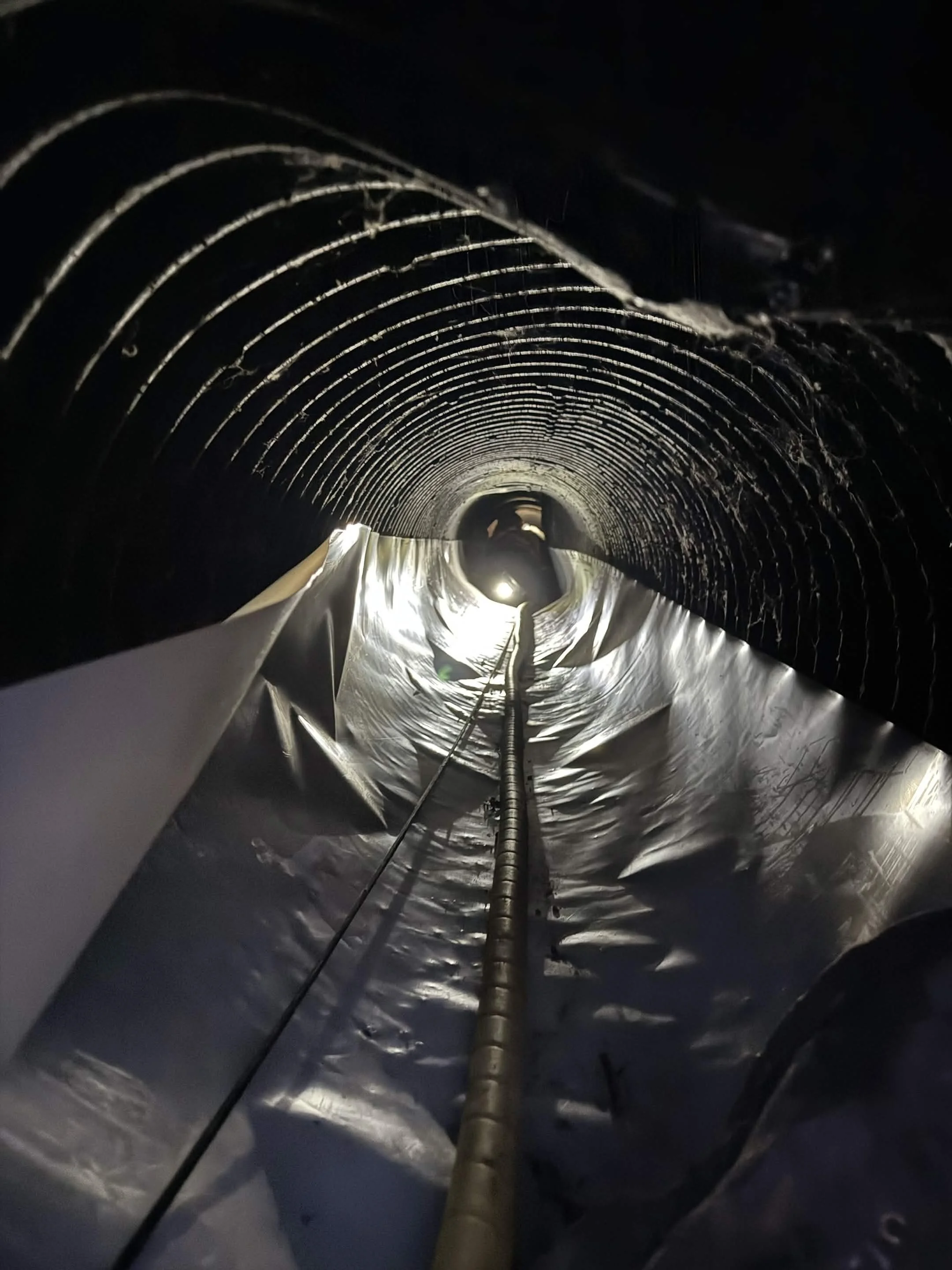 Looking down a long, dark, metallic tunnel with a central support beam and a reflective surface beneath, with the tunnel's curved ceiling made of layered, ribbed metal.