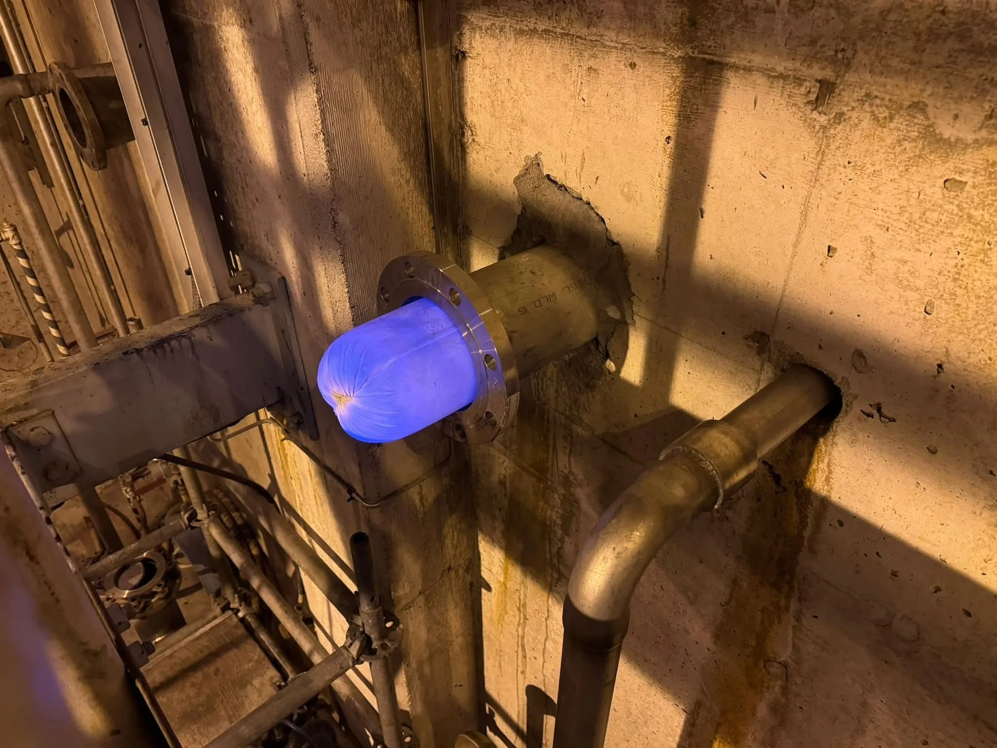 A section of piping and industrial equipment in a facility, with some pipes and a pipe flange emitting blue light.