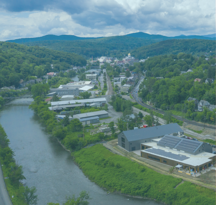 Aerial view of a small town nestled along a winding river, surrounded by lush green hills under a partly cloudy sky.