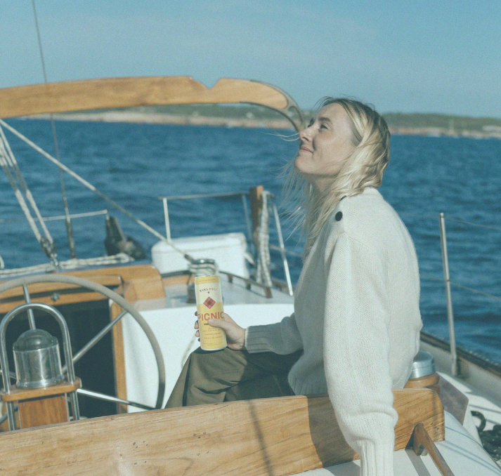 A woman with blonde hair sitting on a boat, holding a bottle of sunscreen, enjoying the sunlight with the ocean in the background.