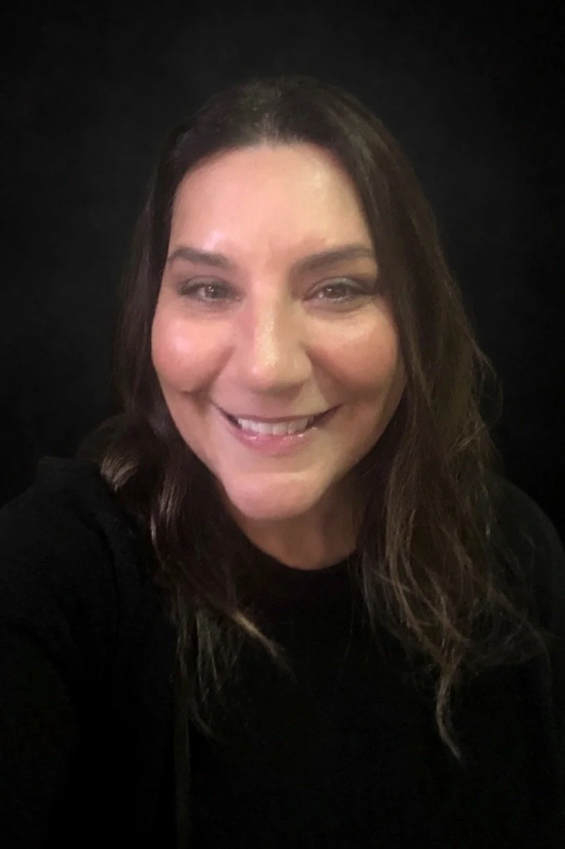 A smiling woman with long brown hair wearing a black top, taking a selfie against a black background.