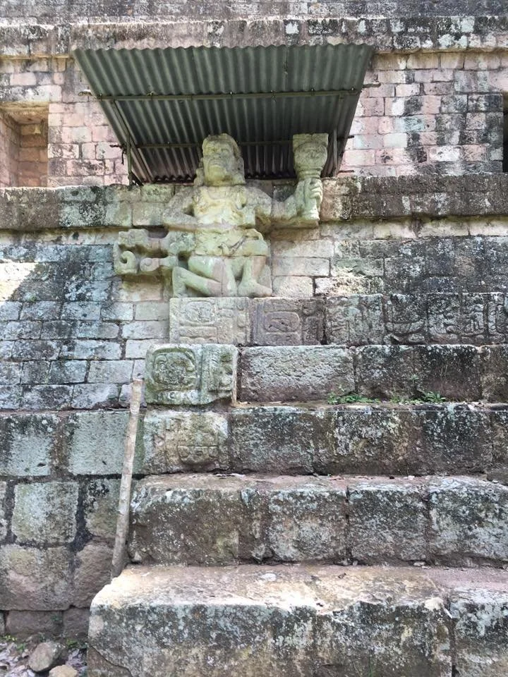 Ancient stone sculpture of a figure with a staff and a second figure, located on a weathered stone staircase beneath a rusted metal roof, with moss and small plants growing on the stones.