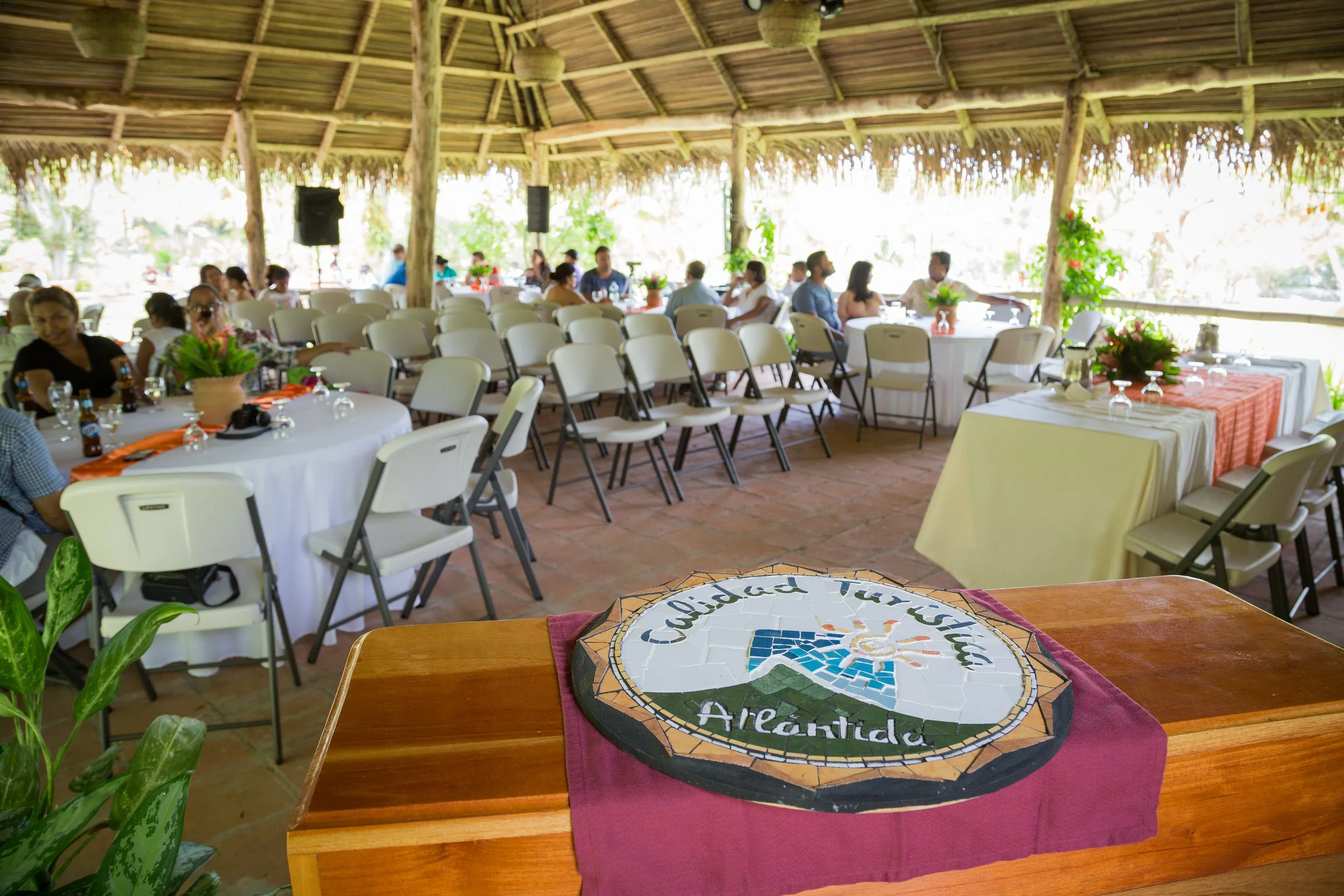 An open-air pavilion with a high thatched roof and wooden beams, decorated with hanging woven baskets and lush green plants. Inside, guests are seated at round tables with white tablecloths, floral centerpieces, glassware, and orange napkins, enjoying a gathering or party.