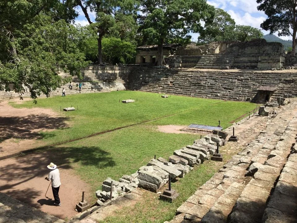 Ancient archaeological site with stone ruins and a grassy area, trees provide shade, few visitors explore the site, and a man wearing a hat walks with a cane.