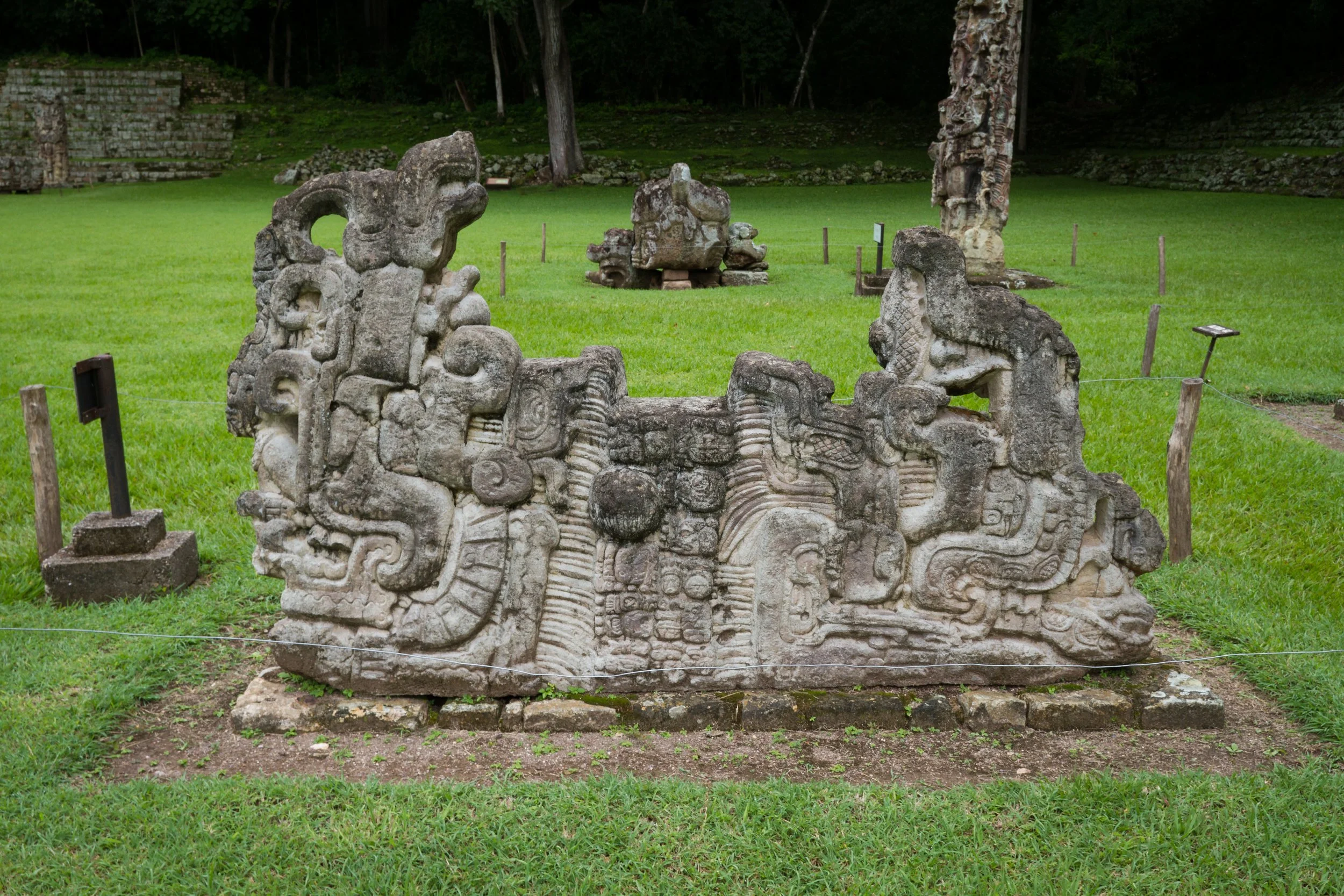 Ancient stone carvings of mythological creatures and figures displayed outdoors on a grassy area, surrounded by a barrier.