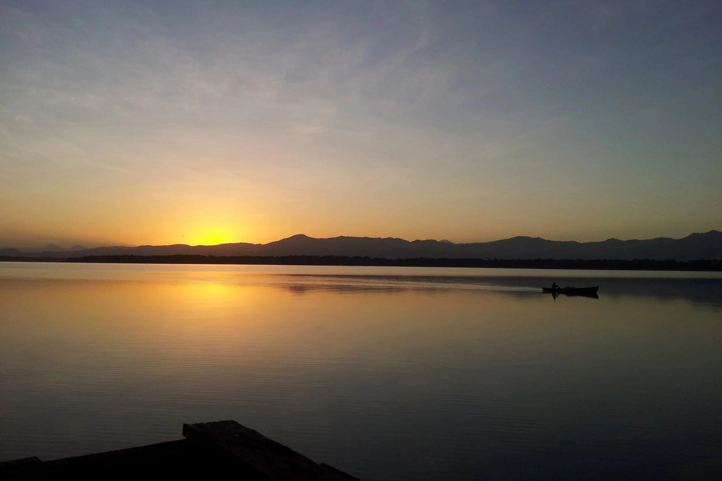 A serene sunset over a calm body of water with mountains in the background. A small boat is floating on the water, and there are wooden logs or structures in the foreground.