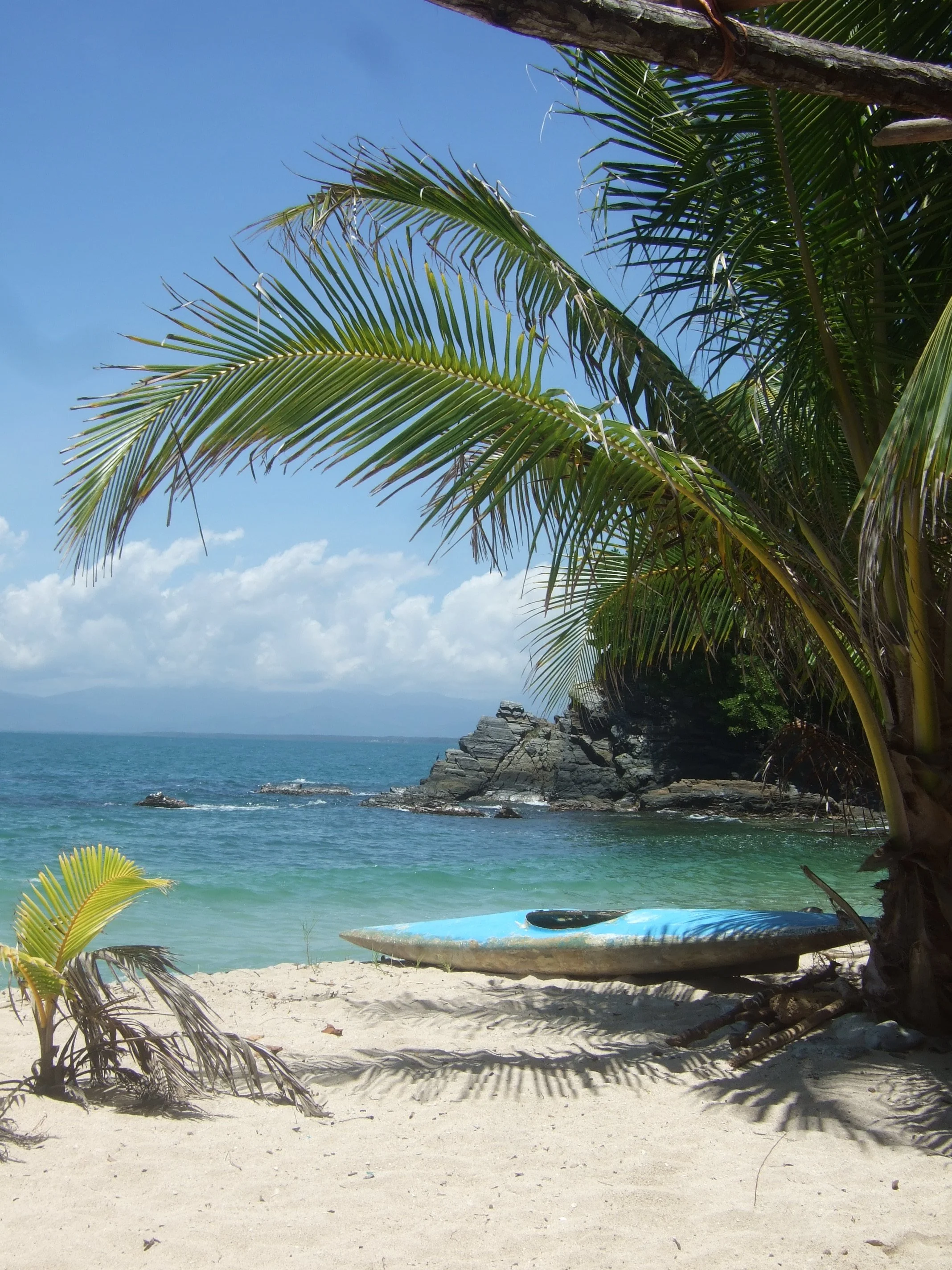 A tropical beach scene with white sand, clear turquoise water, and a rocky coastline. Palm trees with large green fronds shade the beach and a blue surfboard rests on the sand. The sky is mostly clear with some clouds and mountains can be seen in the distance across the water.