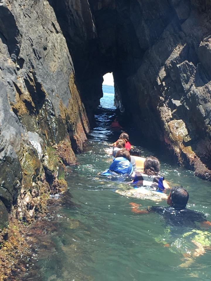 People swimming through a narrow rocky water passage with a natural sea arch in the background.