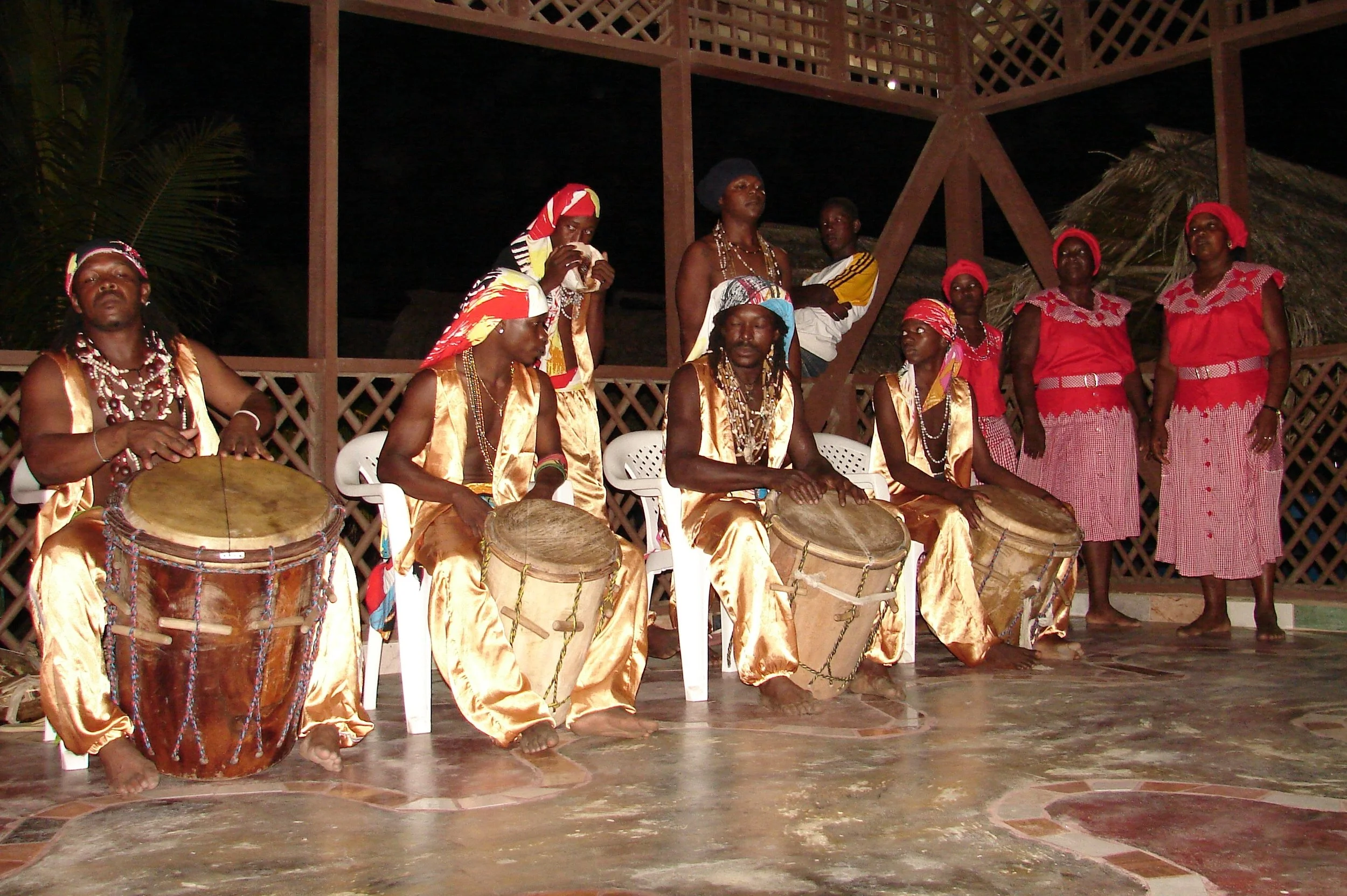 Group of people in traditional clothing performing a cultural dance on stage, with some playing drums, at night under wooden structures and thatched roofing.