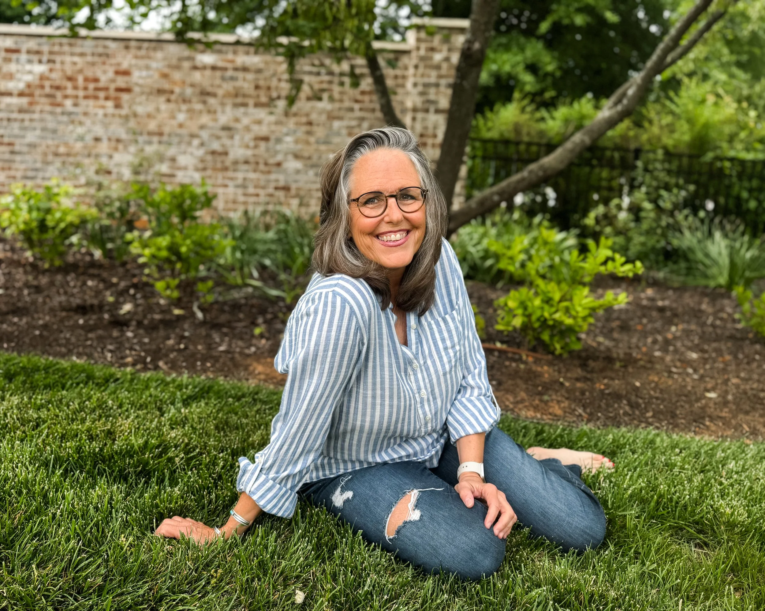 A middle-aged woman with gray hair and glasses smiling and sitting on the grass in a garden with a brick wall, trees, and bushes in the background.