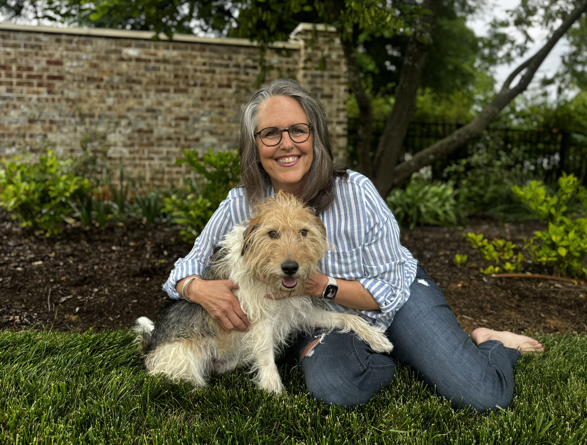 An elderly woman with glasses and gray hair, smiling, sits on the grass holding a scruffy, light-colored dog with a darker tail in her lap, surrounded by green plants and trees, with a brick wall in the background.