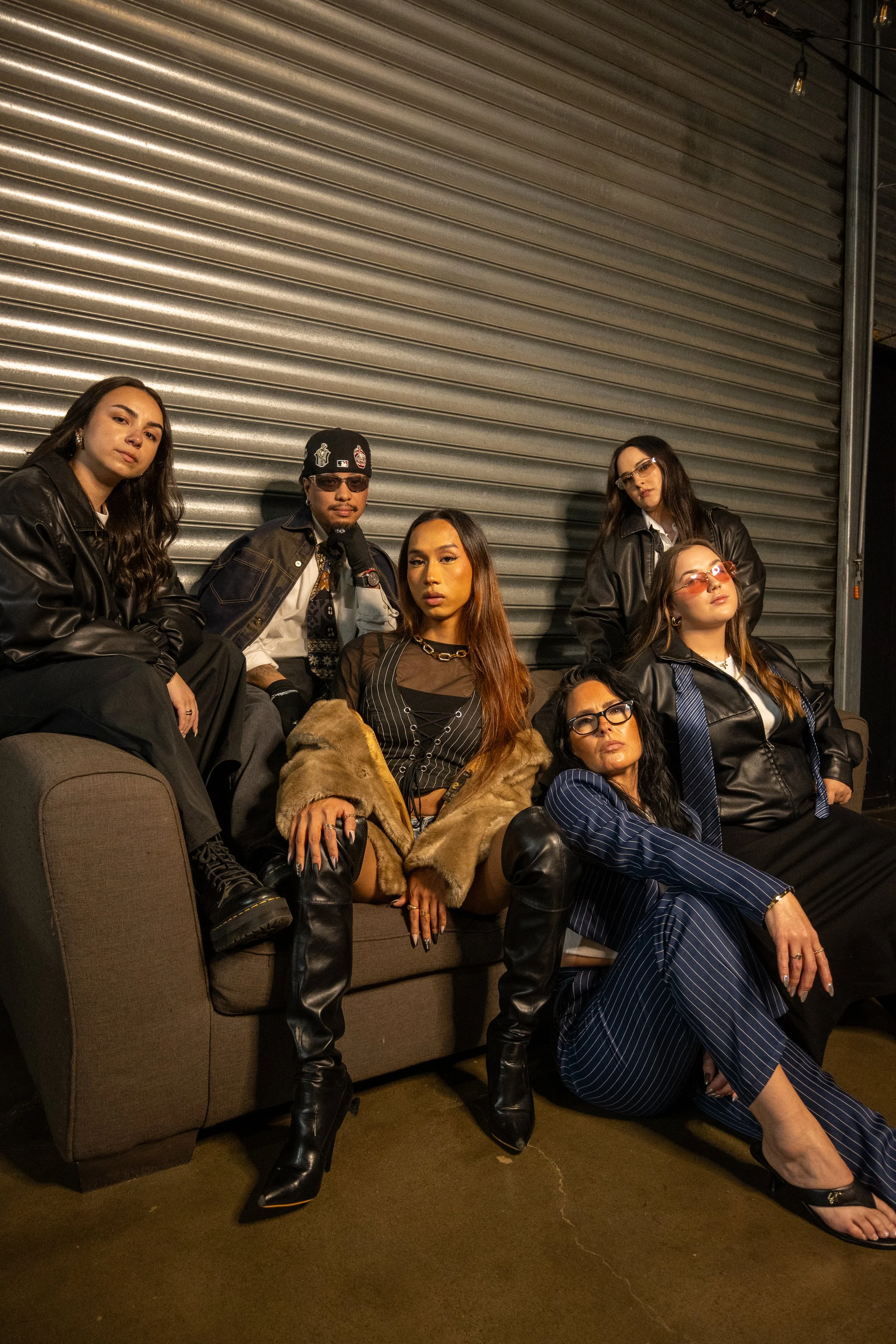 Group of six young adults, five women and one man, posing together in front of a metallic rolling door in an indoor setting. They are dressed in trendy, dark-colored clothing with leather jackets and accessories, and have serious, confident expressions.