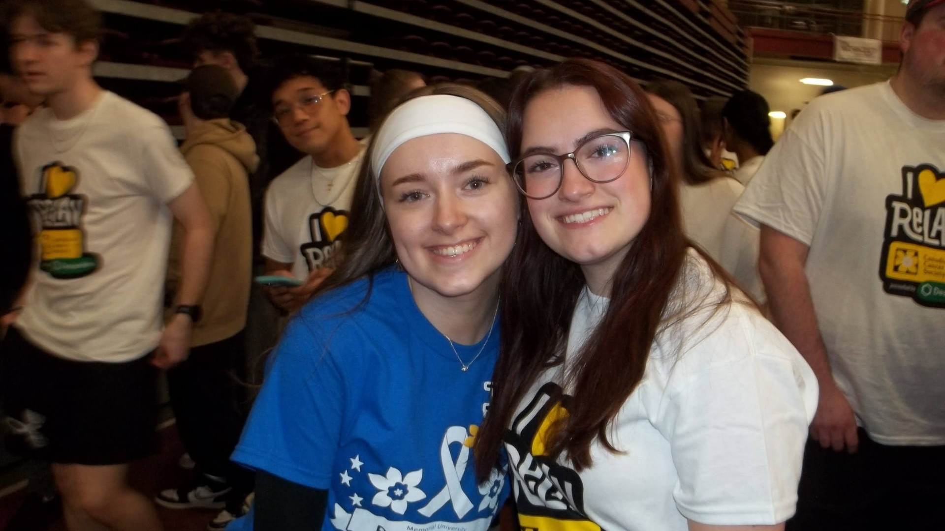 Two young women smiling at the camera, one with long brown hair wearing glasses and a white headband, and the other with long red hair and glasses, at an indoor event with a crowd of young people in the background, many wearing white shirts with a yellow and black logo.