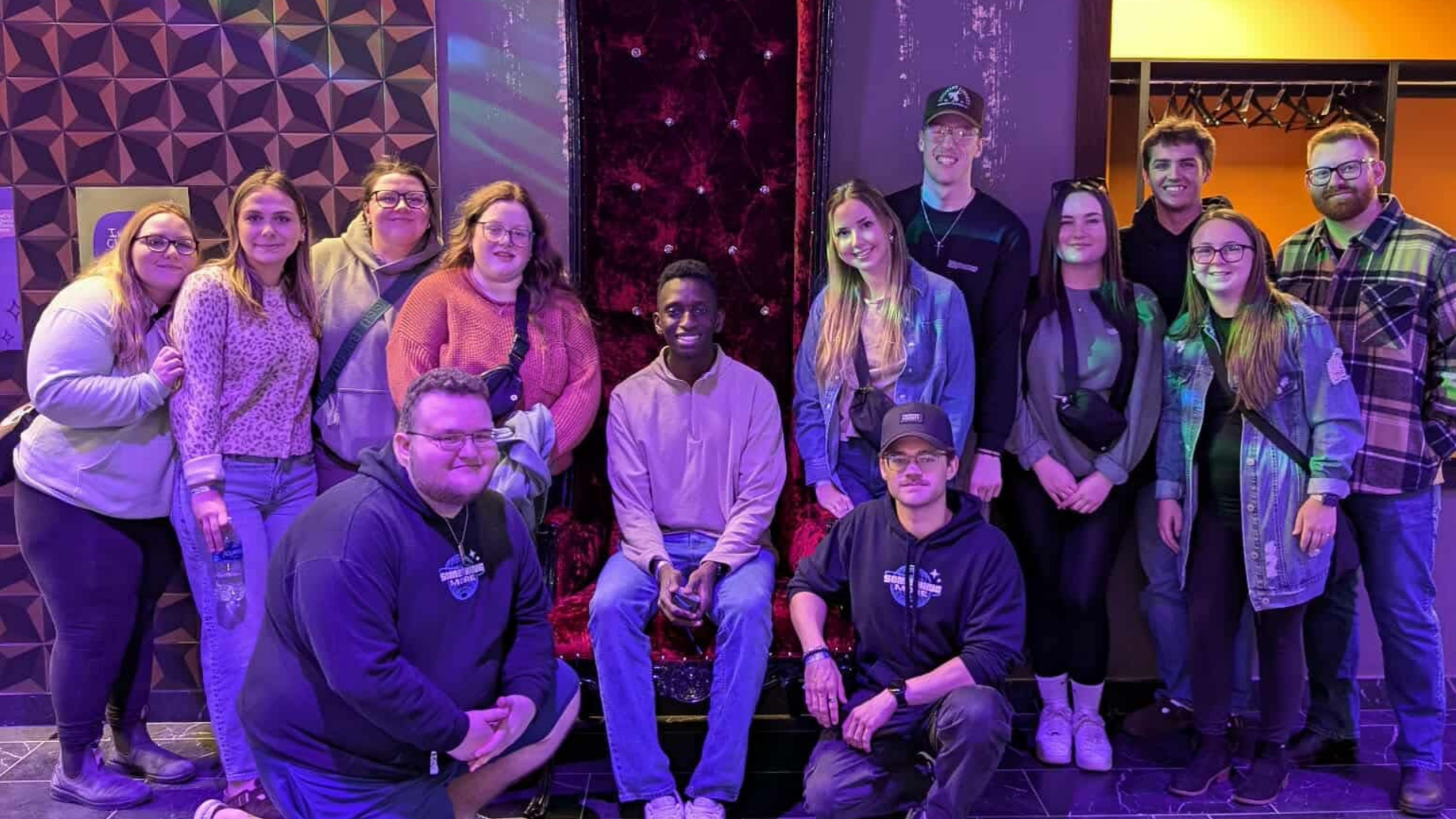 A group of people, including young adults and a young man sitting on a red velvet throne, posing for a photo in a room with purple lighting and geometric wall patterns.