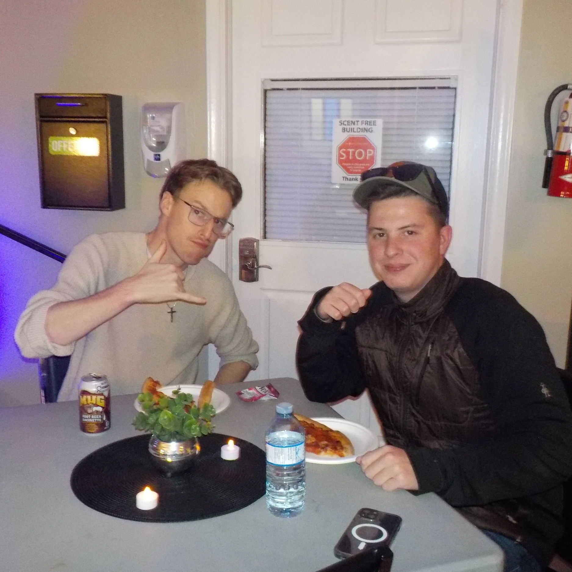 Two young men sitting at a table with pizza, drinks, and a small decorated plant. One is holding his hand in a fist near his chest, the other is making a fist gesture. There is a candle and a bottled water on the table.