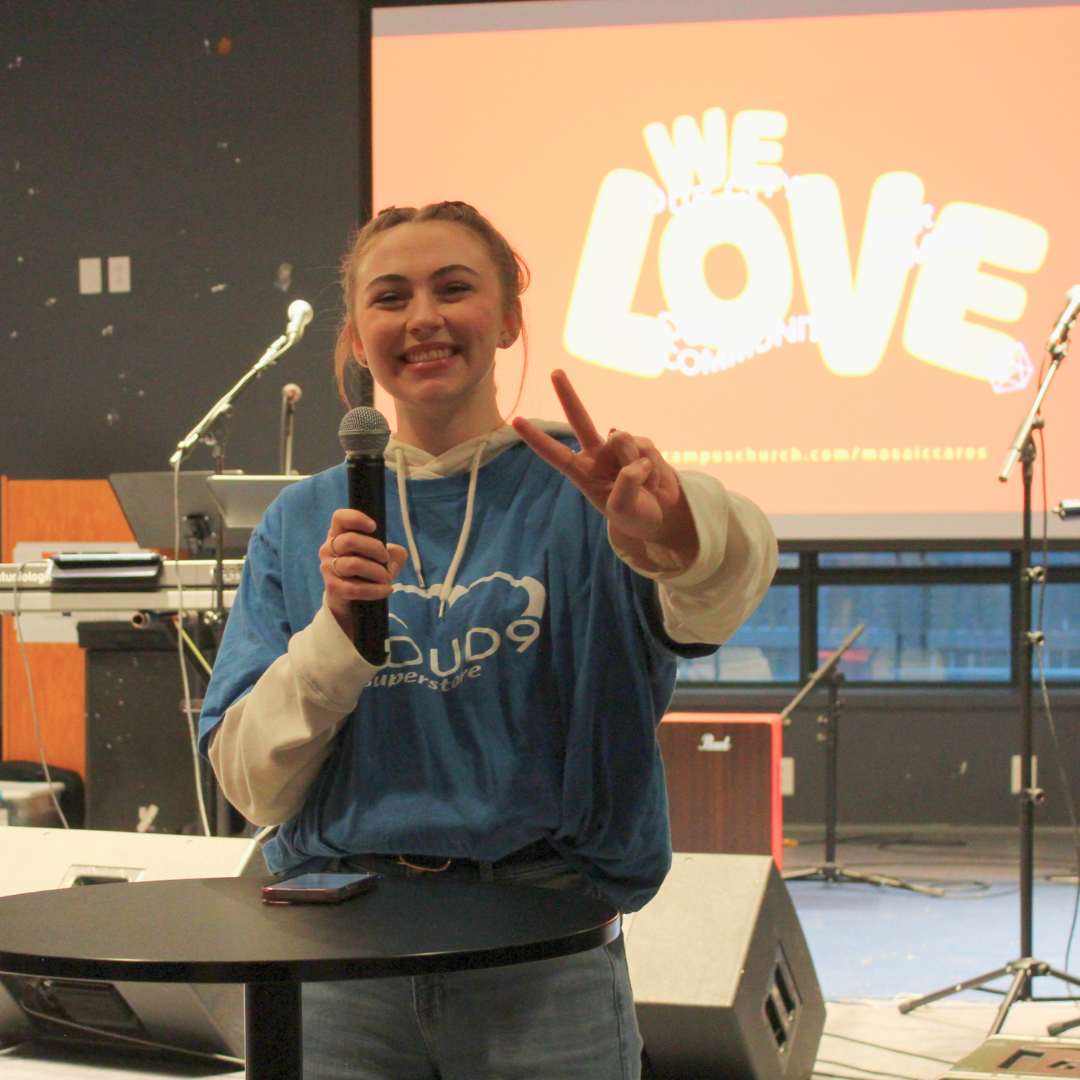 A young woman smiling and making a peace sign with her right hand, holding a microphone in her left hand. She is standing on a stage with musical equipment and a large screen behind her displaying the words 'We Love Community' in bold yellow text on 