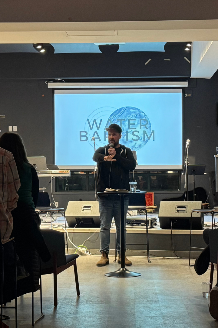 A man in a black hoodie and jeans giving a presentation, standing behind a small round table with a glass of water.