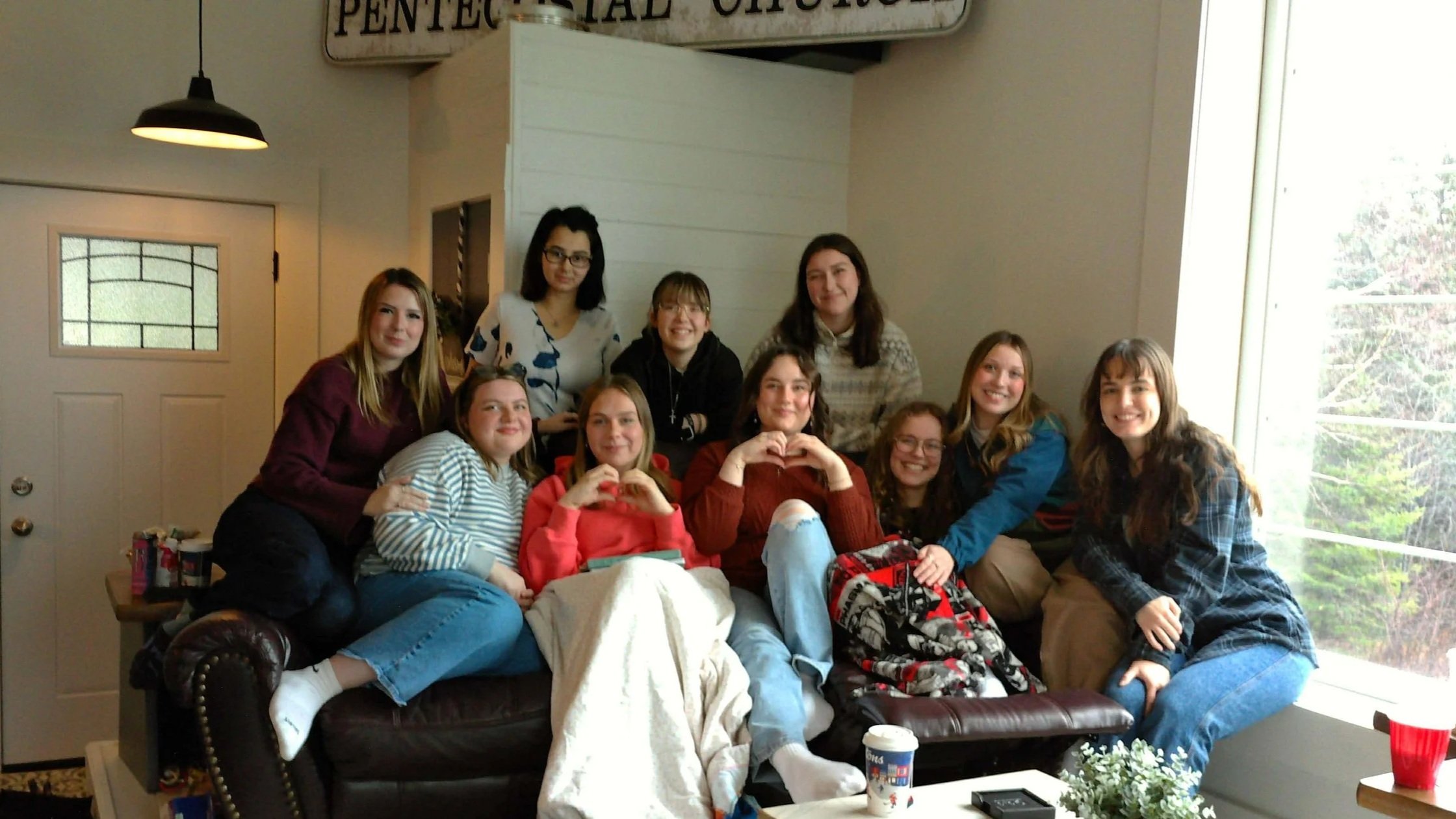 A group of ten young women gathered together on a couch in a cozy indoor setting with a large window in back, showing some greenery outside. They are smiling, some making heart shapes with their hands. The space has a warm, casual atmosphere with som