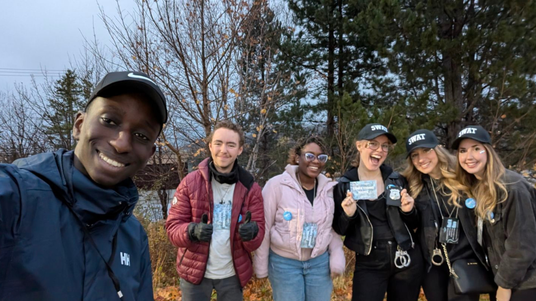 Group of six diverse young adults outdoors during fall, smiling and posing for a selfie. One person is taking the photo, others wear SWAT caps, some hold signs or badges, with trees and cloudy sky in the background.
