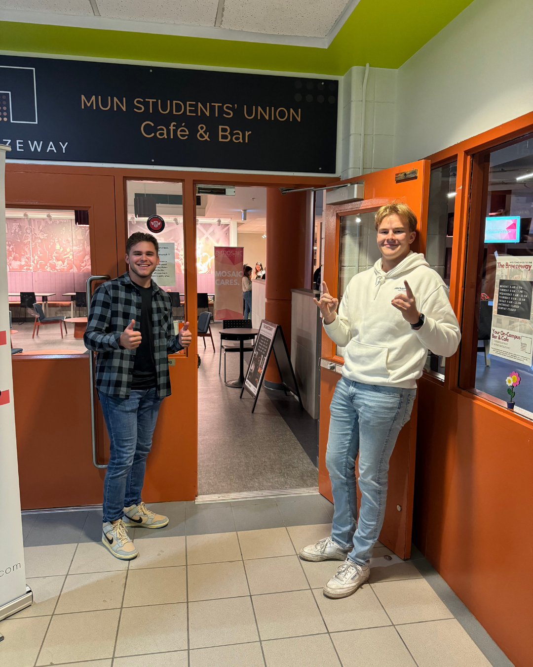 Two young men standing in front of the entrance to the Mun Students' Union Café & Bar, smiling and giving thumbs up gesture.