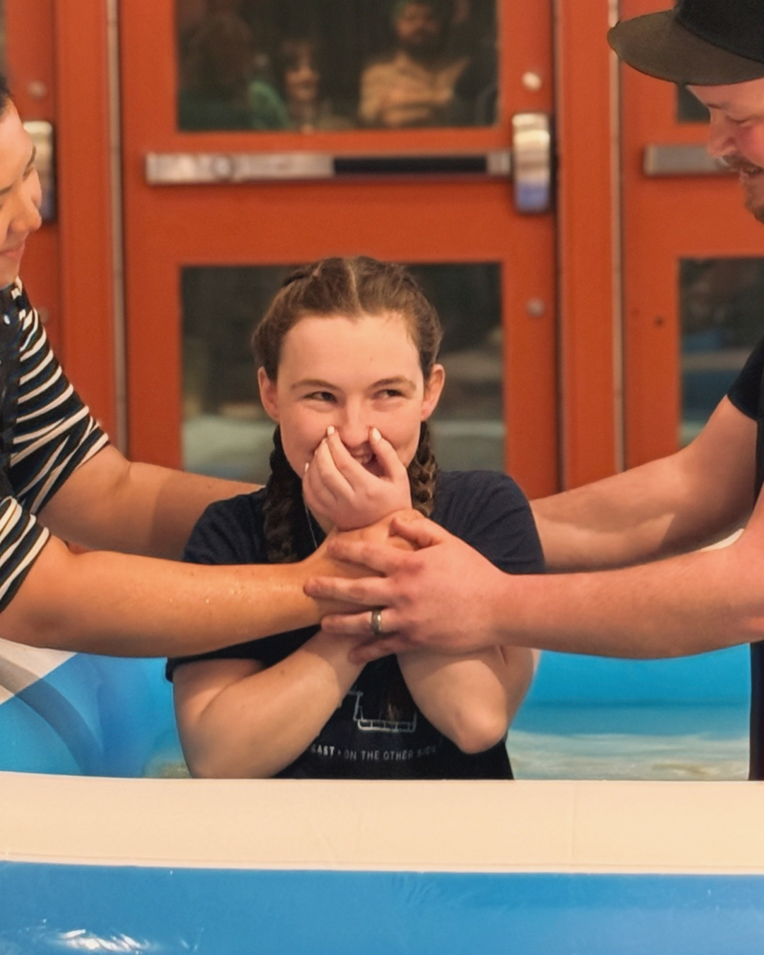 A young girl with braided hair is being baptized in a small pool by two adults, one on each side, holding her hands.