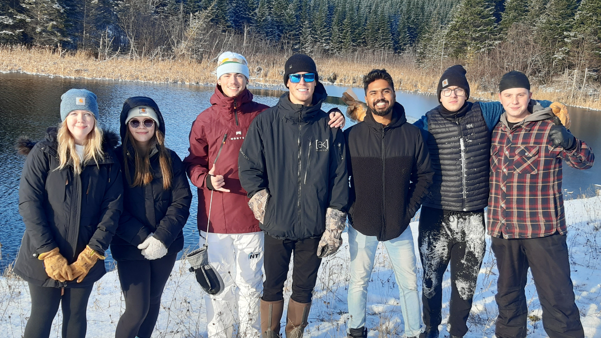 Group of eight friends standing outdoors in winter by a lake, dressed in winter clothing, with snow, trees, and a frozen lake in the background.