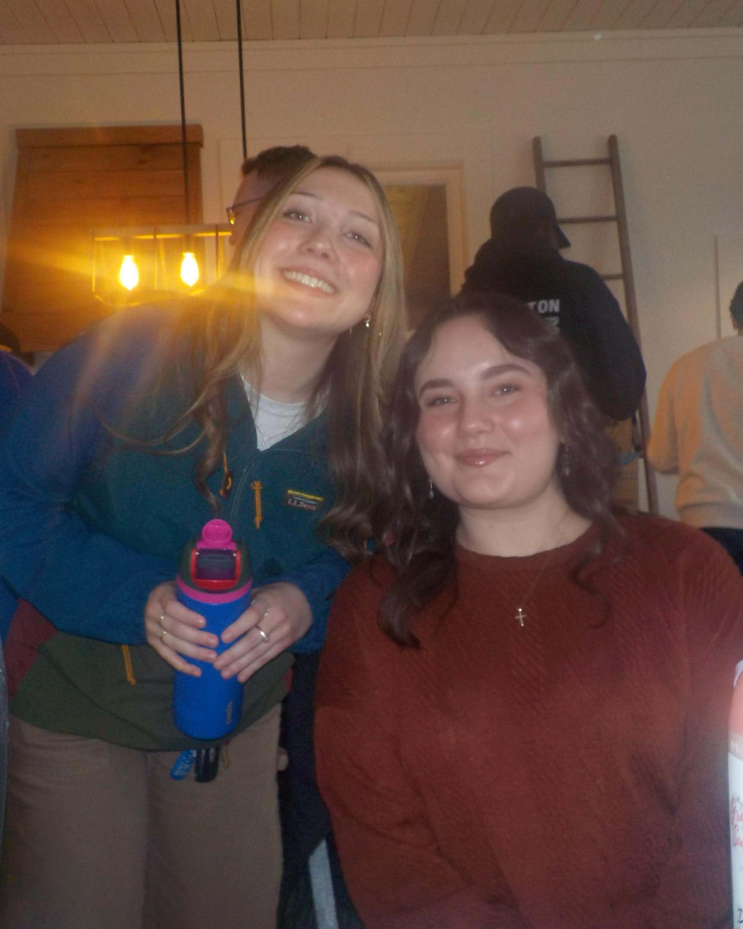 Two women smiling at the camera indoors, with people and a ladder in the background. One woman is holding a water bottle, and there is warm lighting.