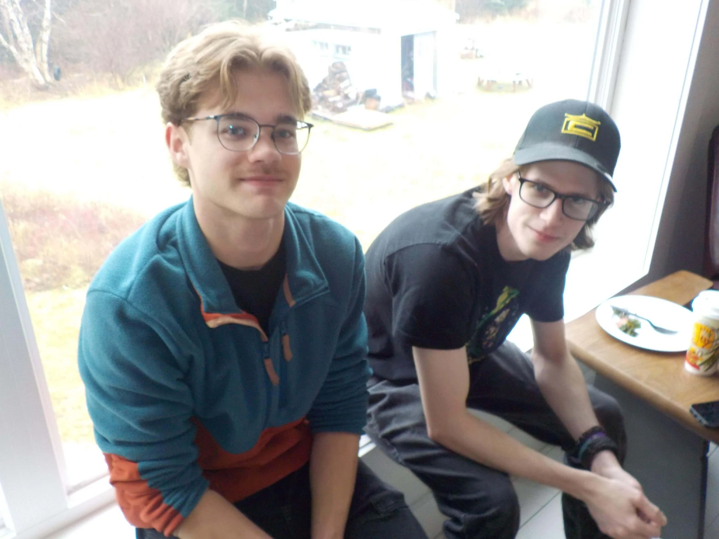 Two young men with glasses sitting at a table near a large window, one with a cap, eating a meal.