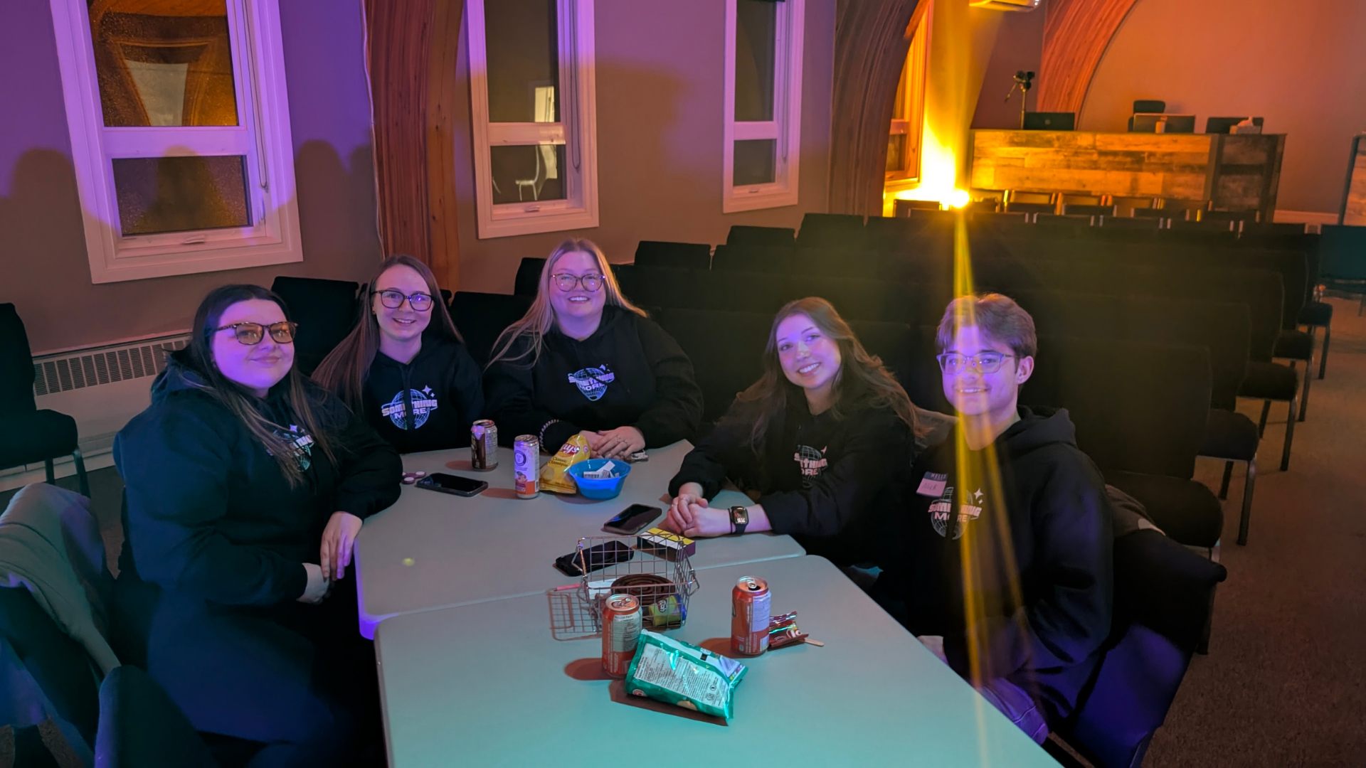 A group of five young people sitting around a table with snacks and drinks in a dimly lit room with purple and yellow lighting, wooden walls, and several empty chairs.