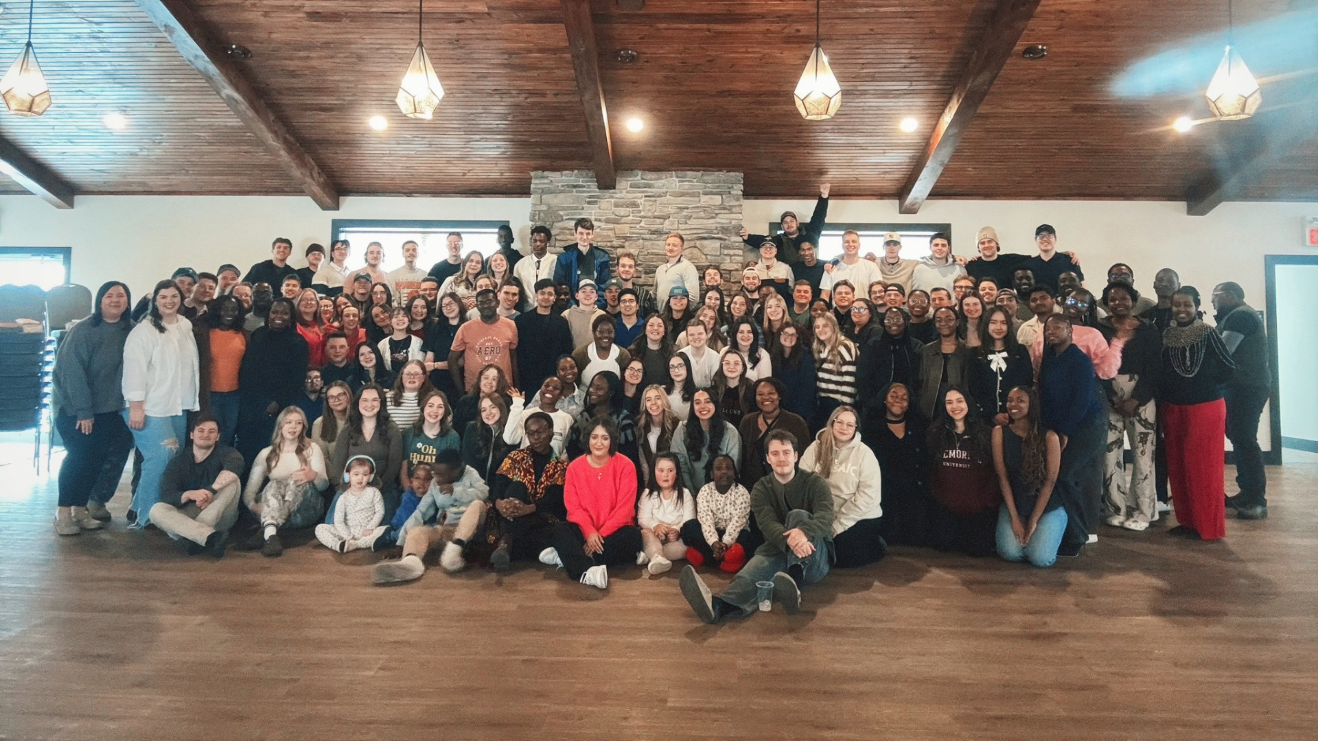 A large diverse group of people gathered in a spacious room with a wooden ceiling and stone accent wall, posing for a group photo. Some are standing, some are sitting or kneeling in the front, and a few are making playful gestures.