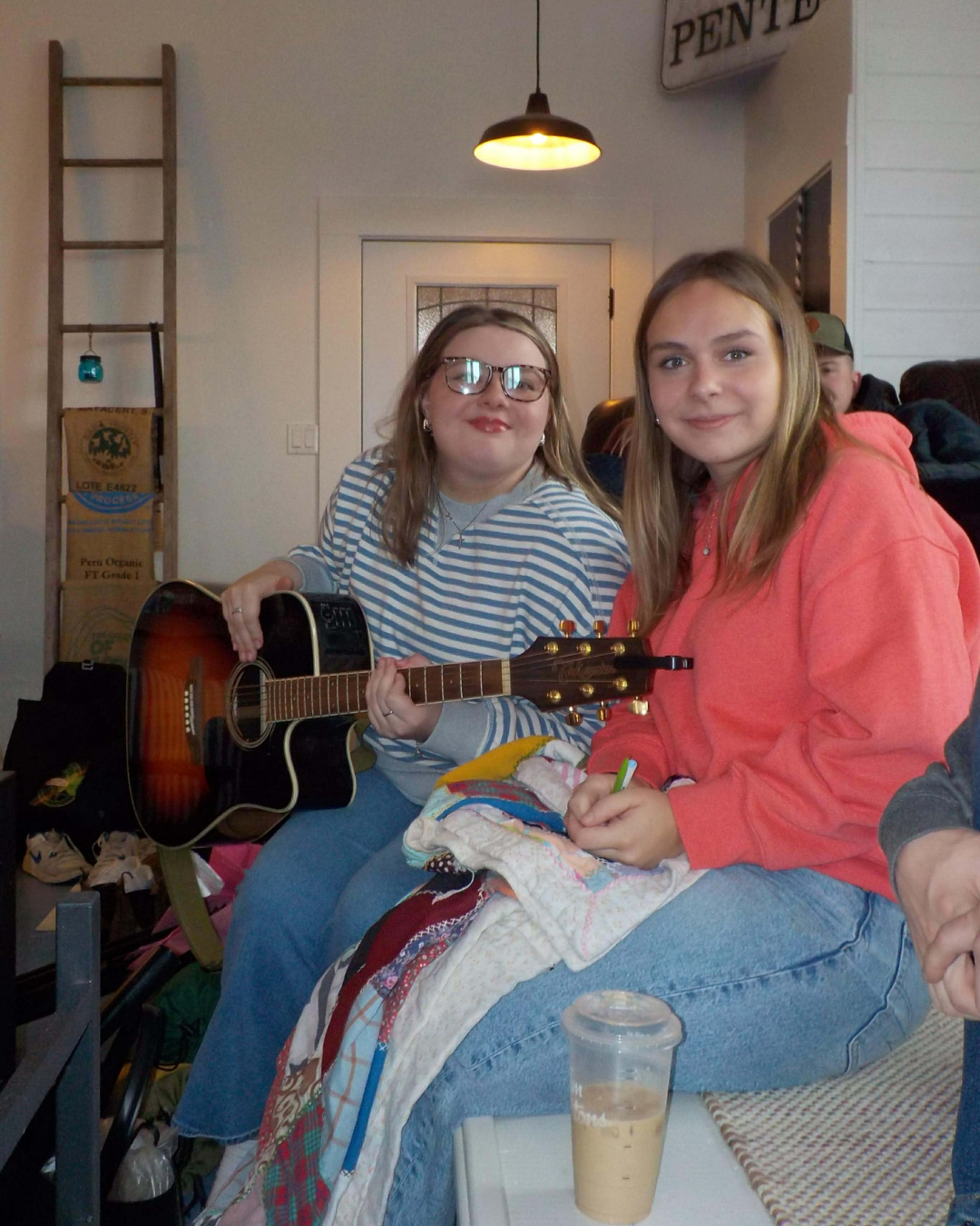 Two young women sitting indoors, one wearing glasses and striped shirt playing guitar, the other wearing pink hoodie smiling, with a coffee drink on the table in front.