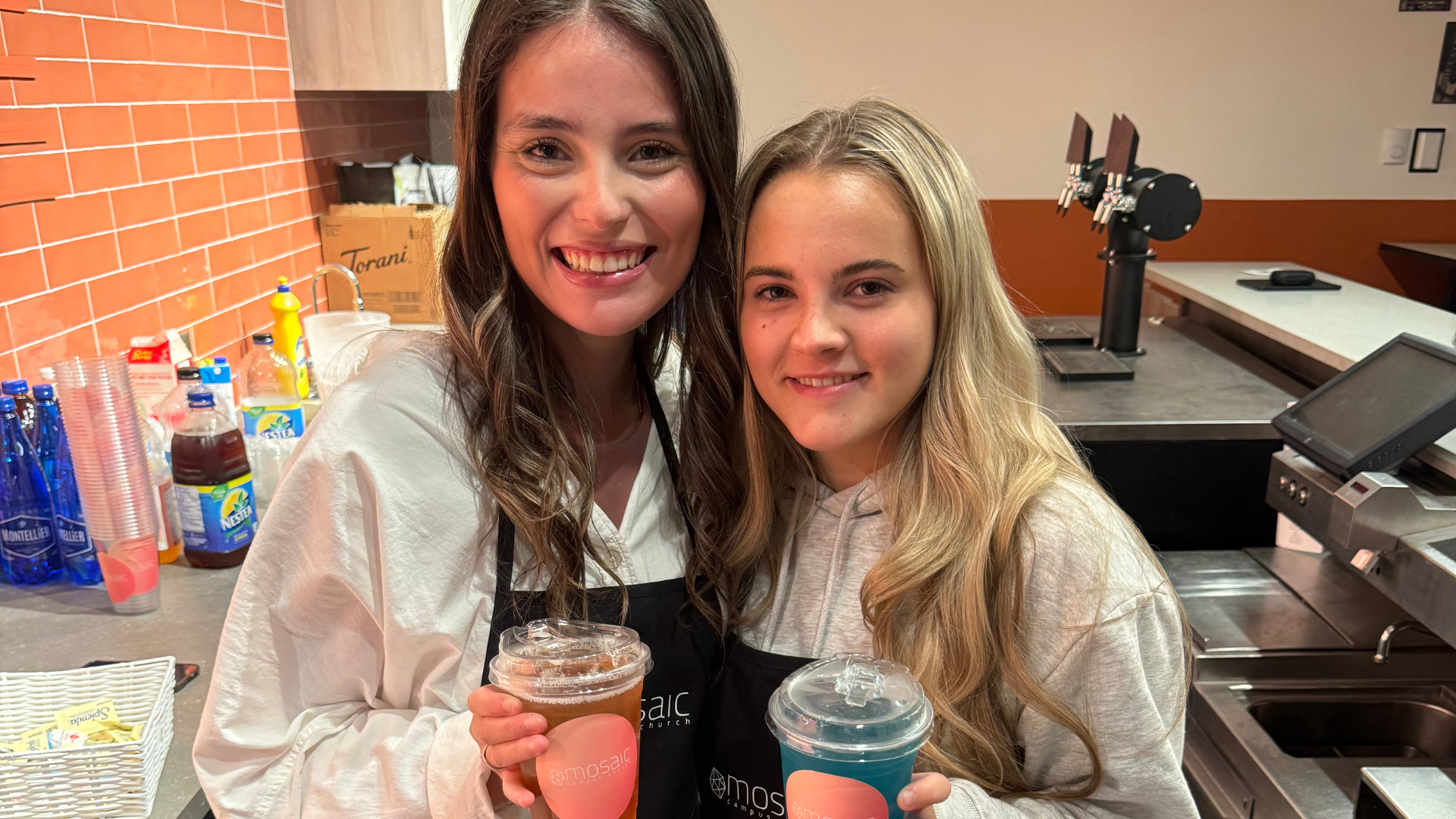 Two young women smiling and holding drinks at a beverage counter in a cafe or restaurant.