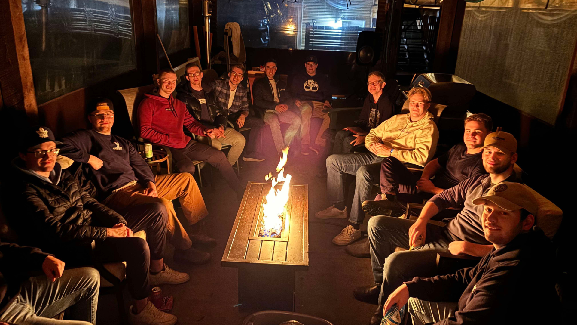 A group of young men sitting around a rectangular fire pit table with a fire burning in the center, in a dimly lit indoor space, smiling at the camera.