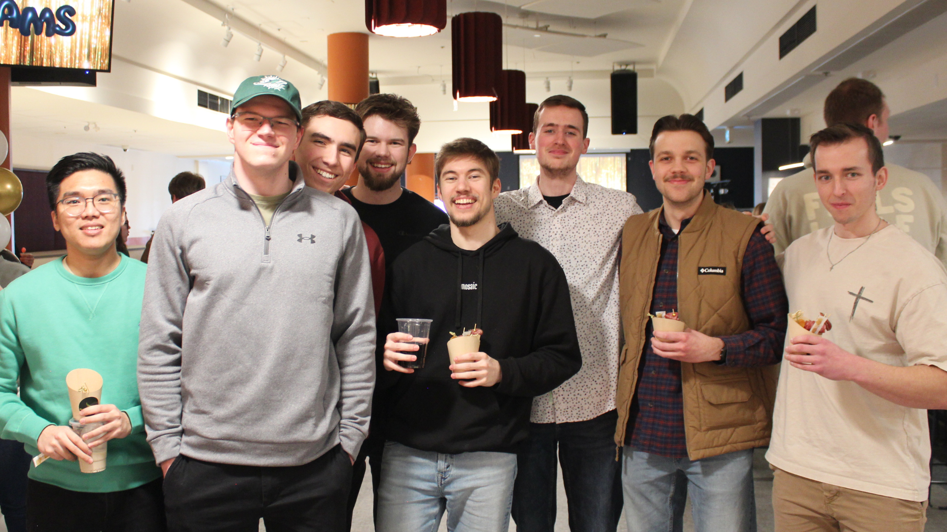 A group of nine young men standing together indoors, smiling at the camera. Some are holding drinks and desserts, and the background shows a modern party or event setting.